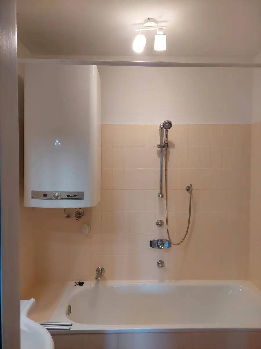 Badezimmer mit Badewanne, Duscharmatur und einem an der Wand montierten Warmwasserbereiter.