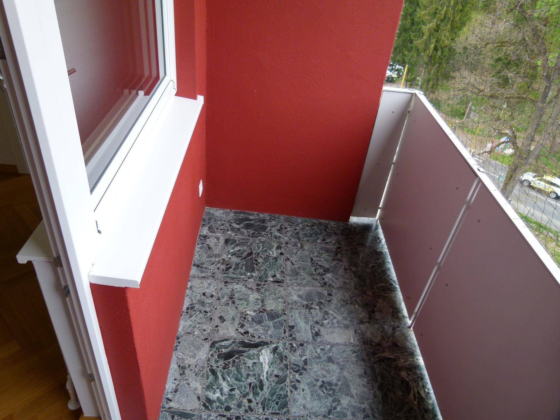 Kleiner Balkon mit dunkelgrünen Fliesen und roter Wand, ideal für eine Person.