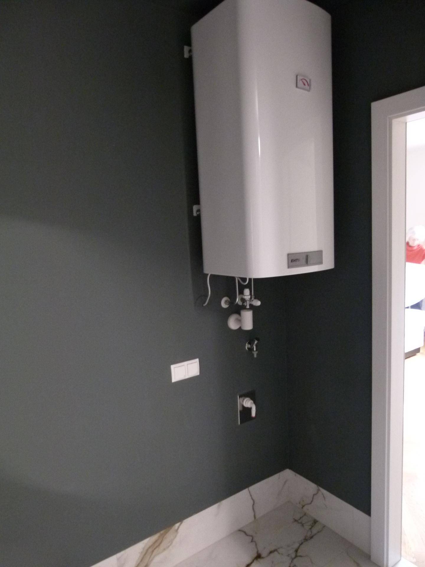 Wandmontierter Warmwasserbereiter in einem Raum mit dunklen Wänden und Marmorfliesen.