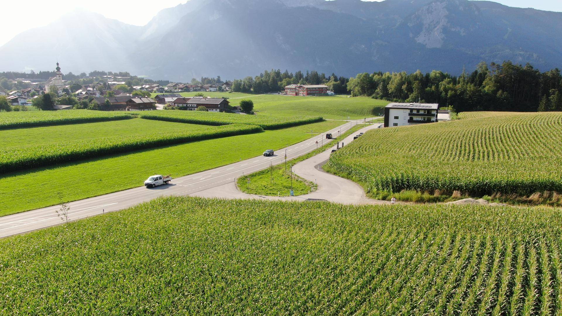 Sonnige Luftaufnahme einer Landstraße, die durch Maisfelder führt, mit einem Dorf und Bergen im Hintergrund.