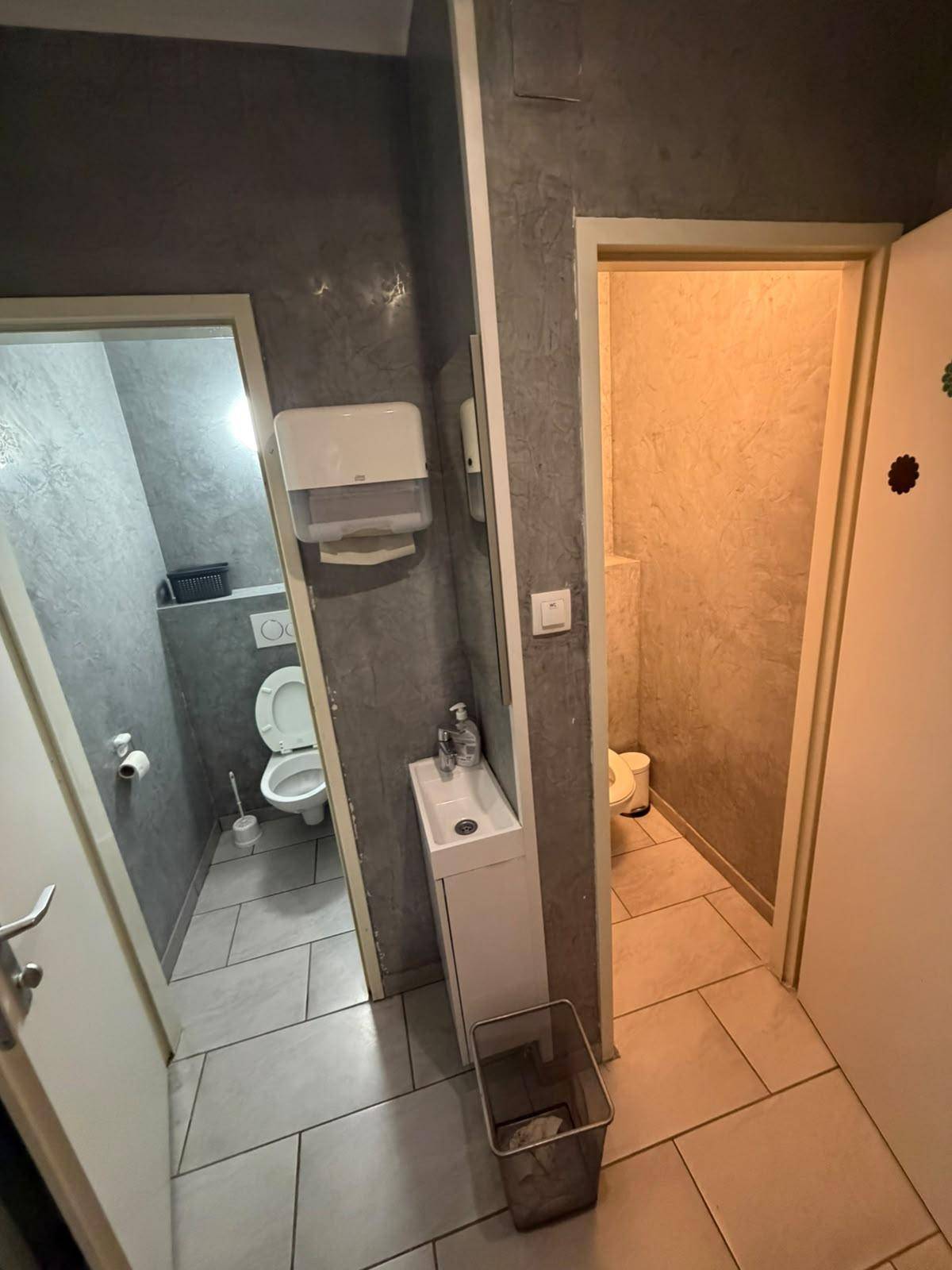 Zwei separate Toilettenräume mit einem gemeinsamen Waschbeckenbereich, alle mit grauen Wänden und Fliesenboden.
