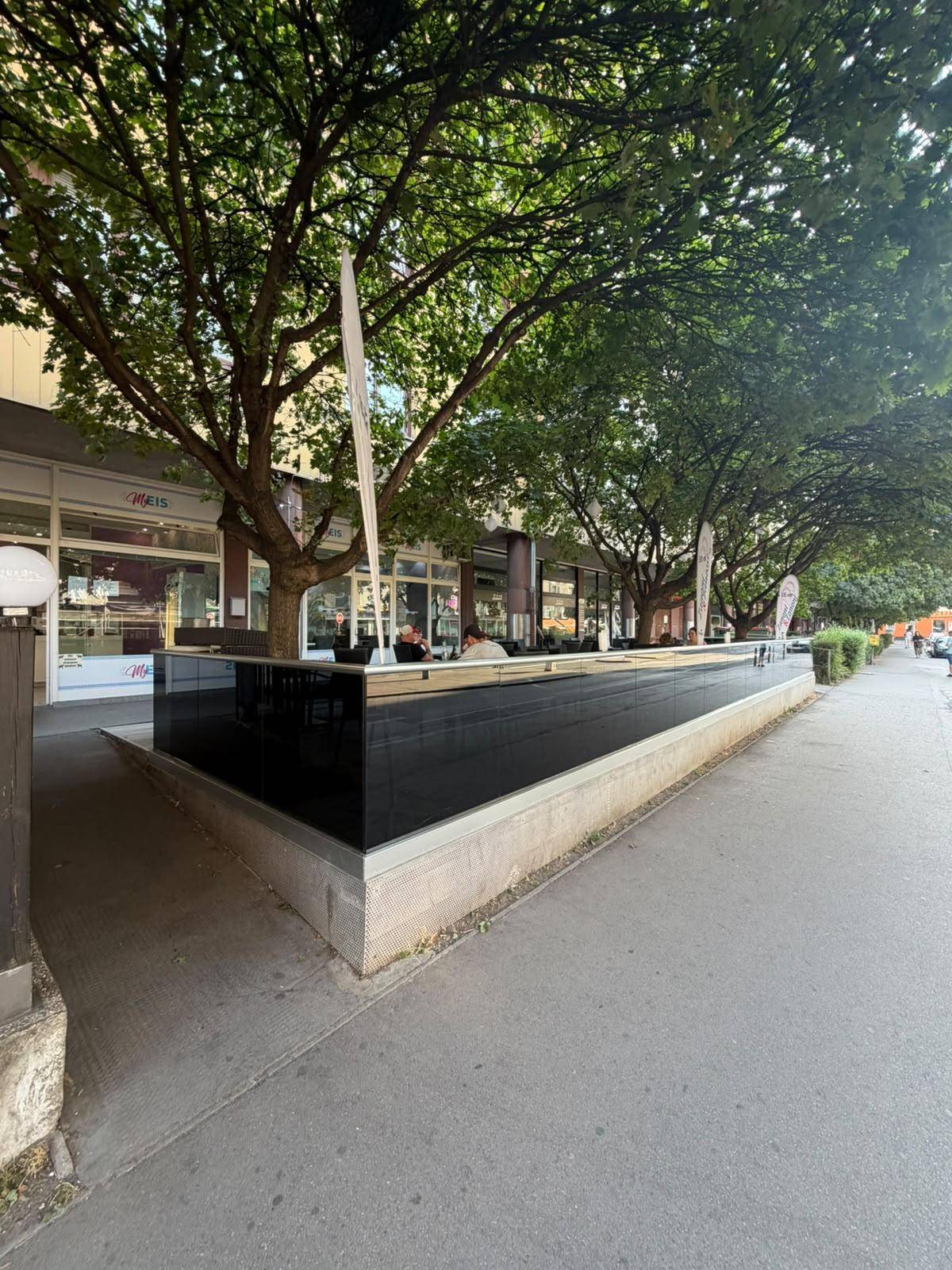 Eine weitere Ansicht der Straße mit üppigen Bäumen, einem langen schwarzen Pflanzgefäß und einem Café mit Gästen im Freien.
