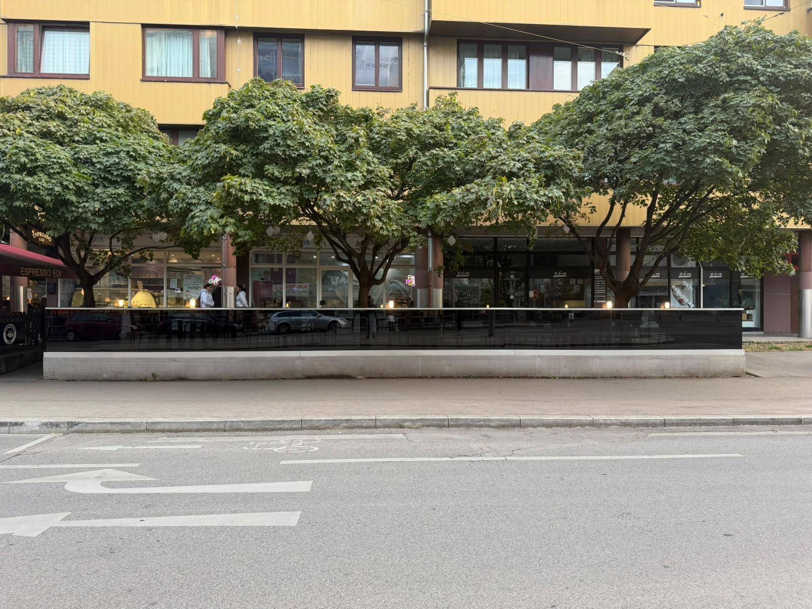 Gebäudefassade mit großen Fenstern im Erdgeschoss, umgeben von Bäumen und einer belebten Straße.