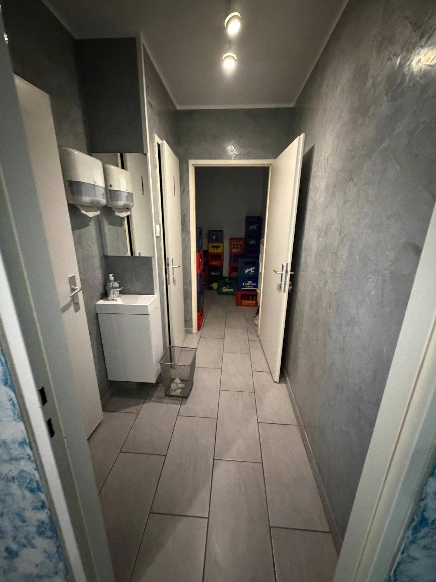 Flur mit grauen Wänden und Fliesenboden, der zu einem Lagerraum und den Toiletten führt.