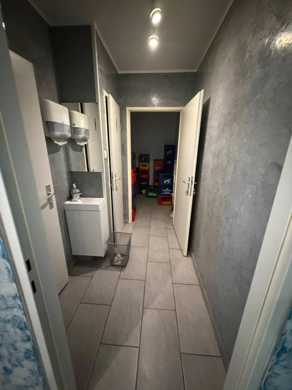 Ein gefliester Flur mit grauen Wänden, der zu einem Lagerraum führt, mit den Toiletten auf der linken Seite.