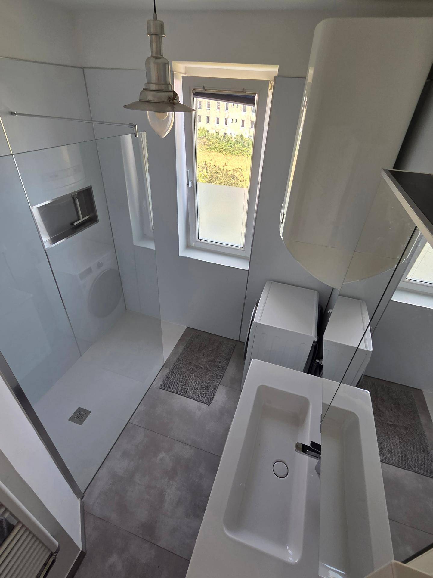 Helles Badezimmer mit begehbarer Dusche, Waschbecken und Waschmaschine unter dem Fenster.