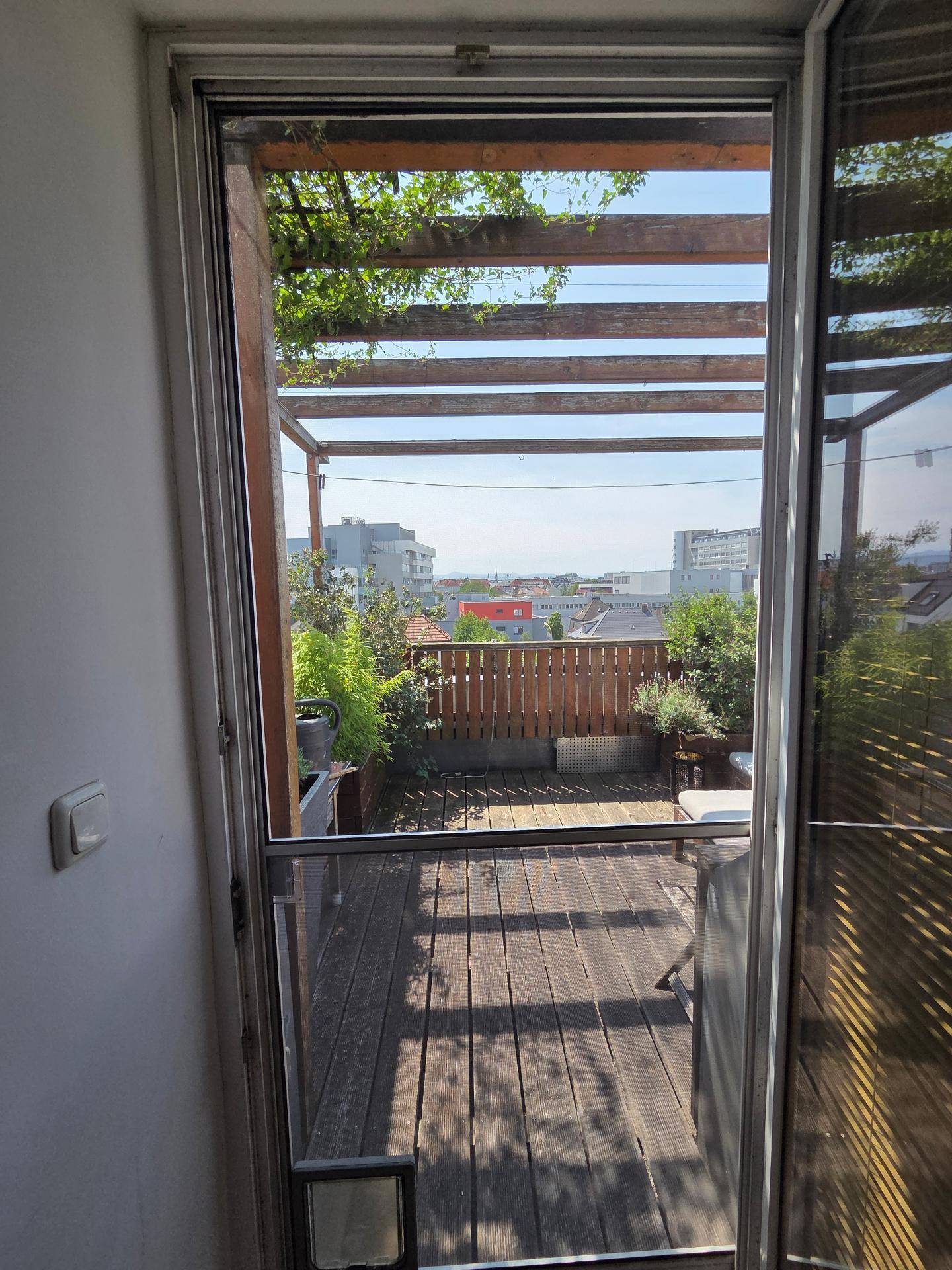 Blick von innen auf eine sonnige Holzterrasse mit Pergola und urbaner Fernsicht.