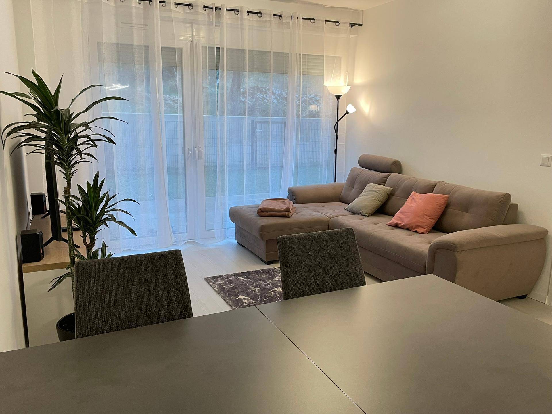 Heller Wohnbereich mit beigem Ecksofa und großen Fenstern zum Außenbereich.