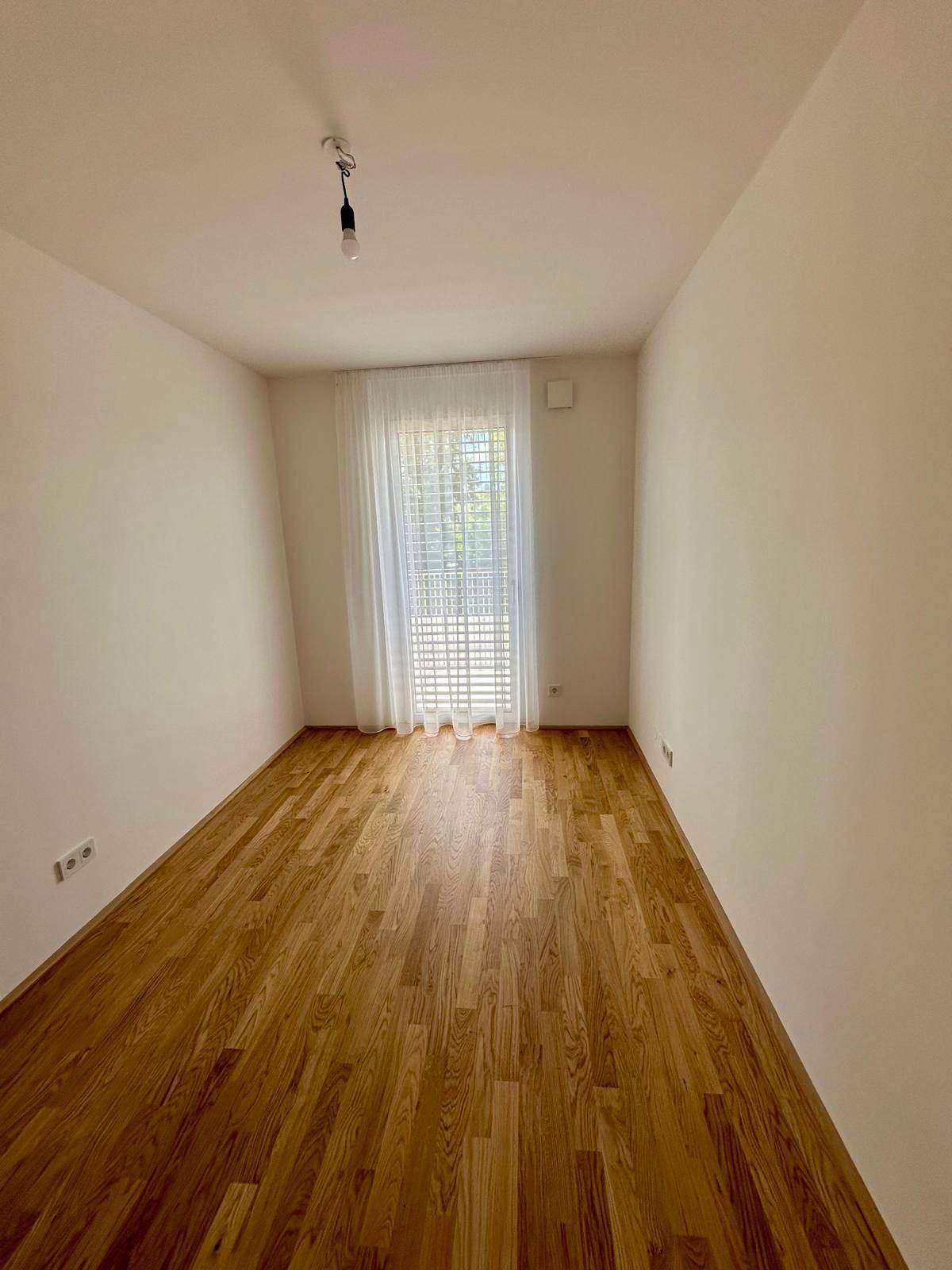 Helles Zimmer mit warmem Holzparkettboden und großem Fenster, ideal als Schlaf- oder Arbeitszimmer.