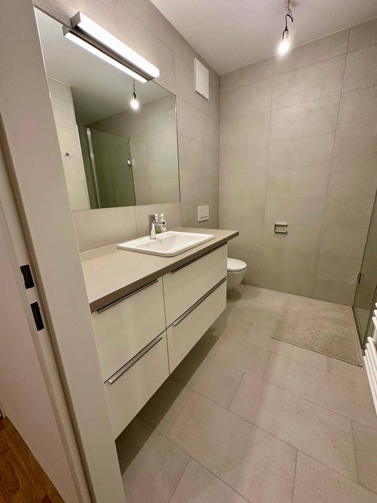 Modernes Badezimmer mit großem Waschtisch, integriertem Spiegel und hellgrauen Fliesen an Wänden und Boden.