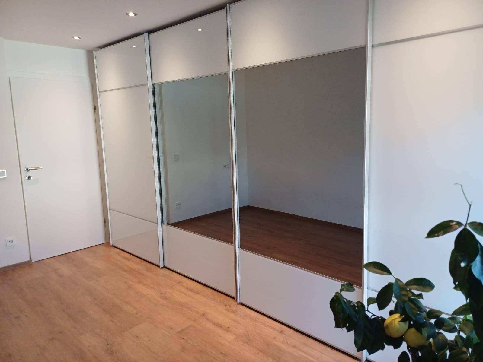 Raum mit hellem Holzfußboden und einer Wand aus weißen Einbauschränken mit Spiegelfronten.