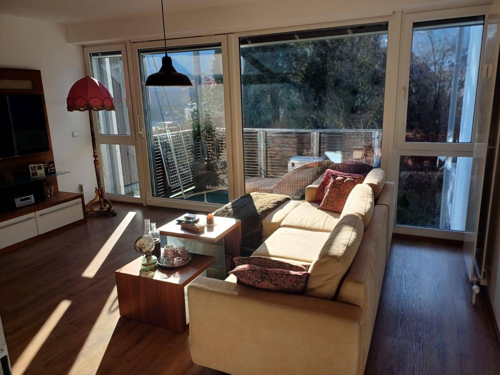 Gemütliches Wohnzimmer mit beigem Sofa, Holztischen und viel Tageslicht durch große Fenster.