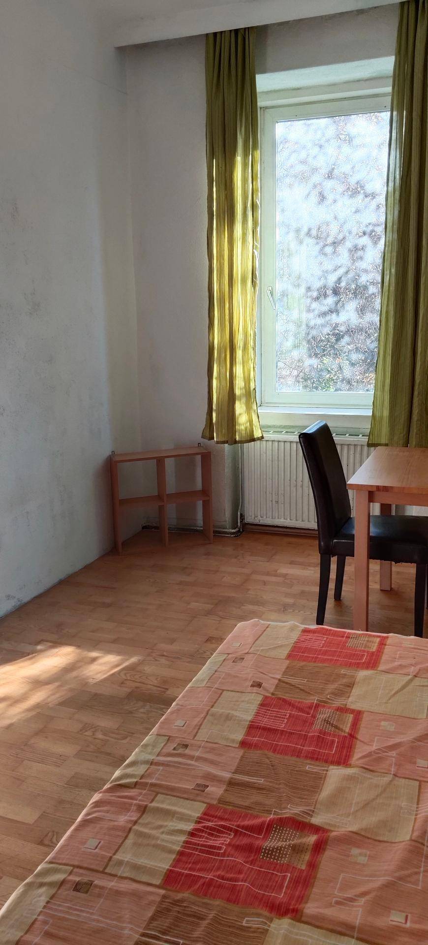 Helles Schlafzimmer mit Holzboden, Fenster und grünen Vorhängen, ausgestattet mit Bett und Schreibtisch.