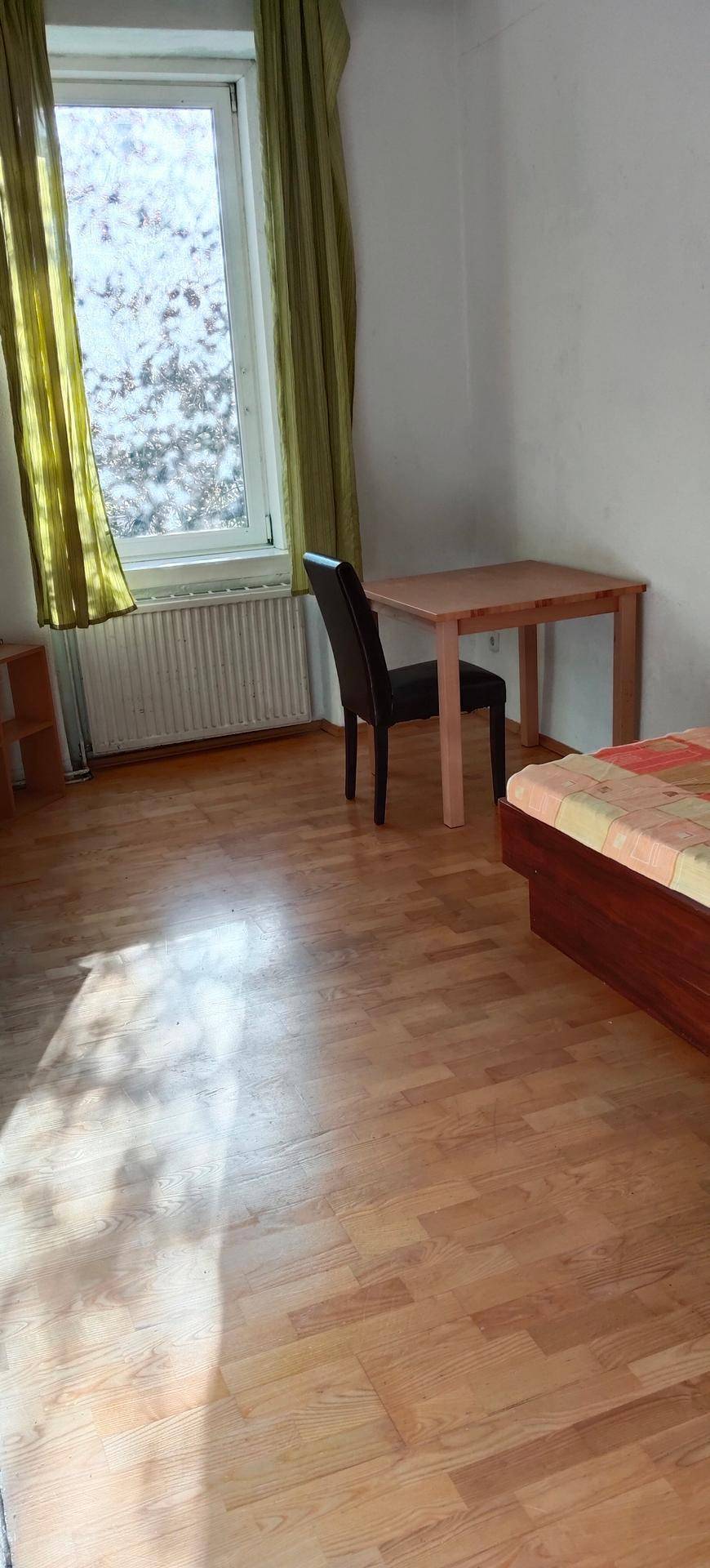 Lichtdurchflutetes Schlafzimmer mit Holzboden, großem Fenster und einfacher Möblierung.