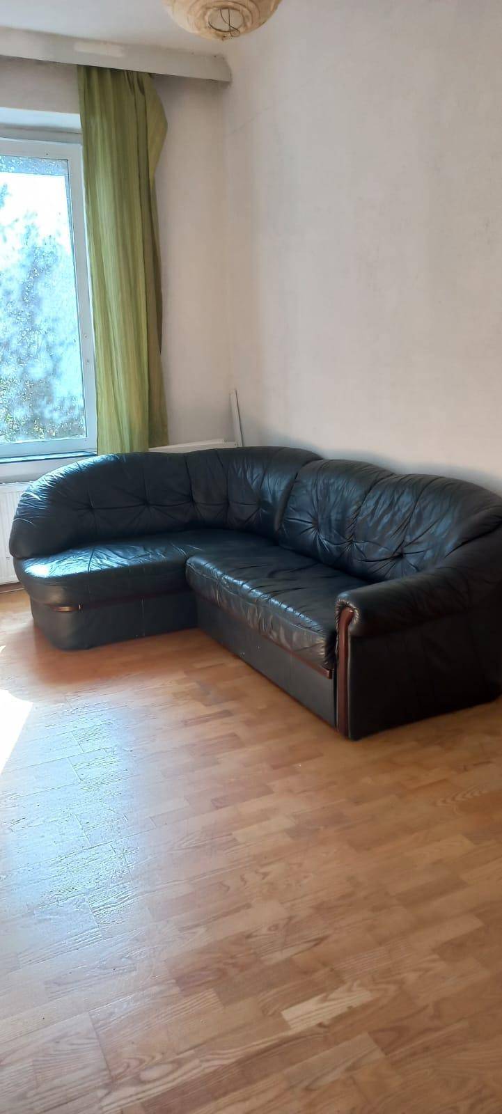 Gemütliches Wohnzimmer mit großem schwarzem Ledersofa, Holzboden und Fenster mit grünen Vorhängen.