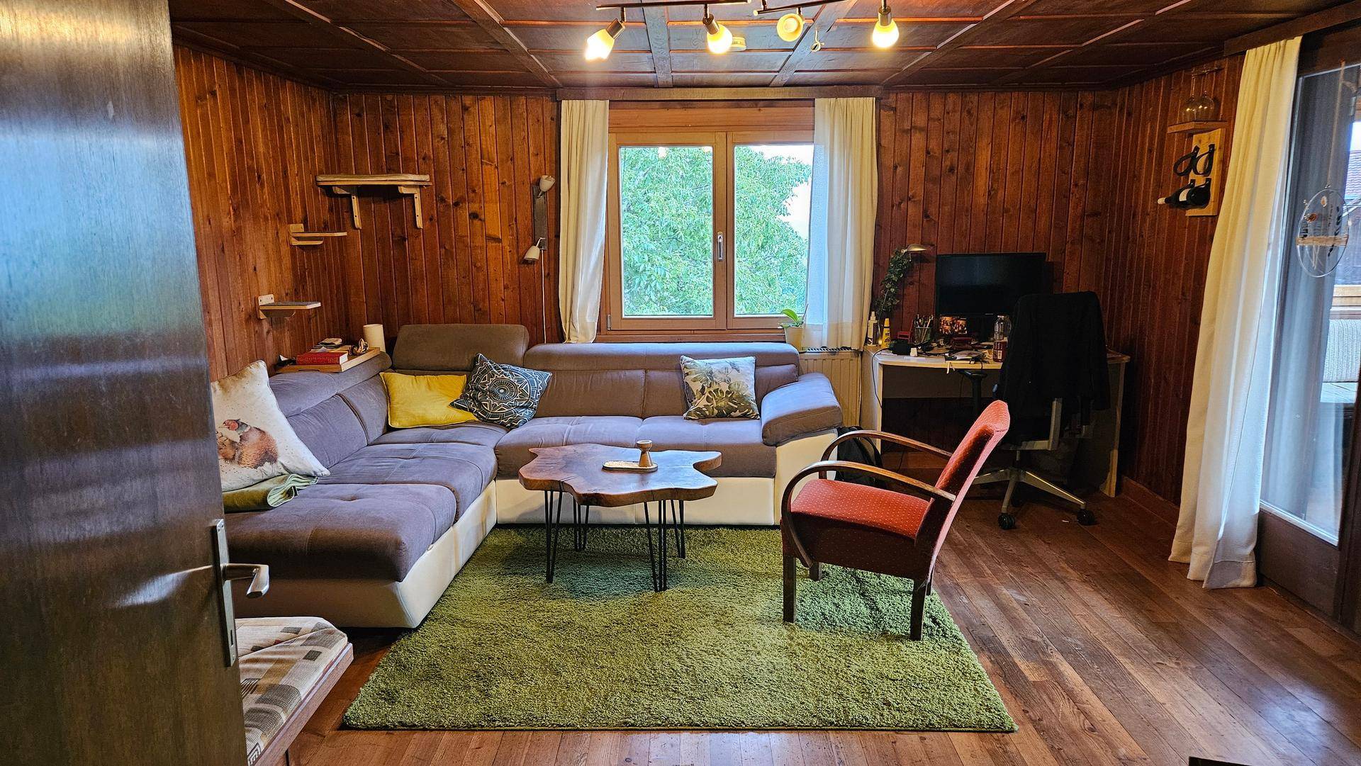 Gemütliches Wohnzimmer mit Holztäfelung, großem Sofa, grünem Teppich und Arbeitsbereich.