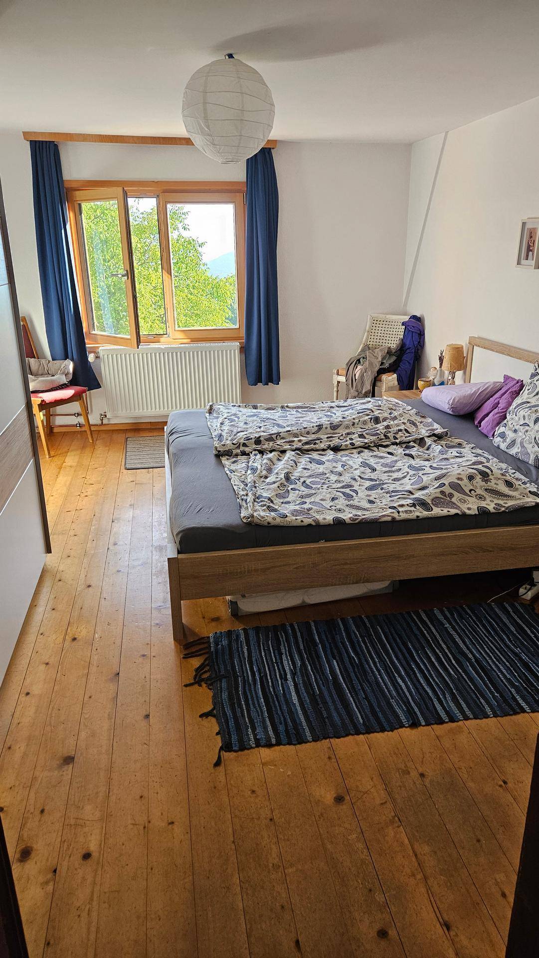 Helles Schlafzimmer mit Holzdielenboden, Doppelbett und Fenster mit blauem Vorhang.