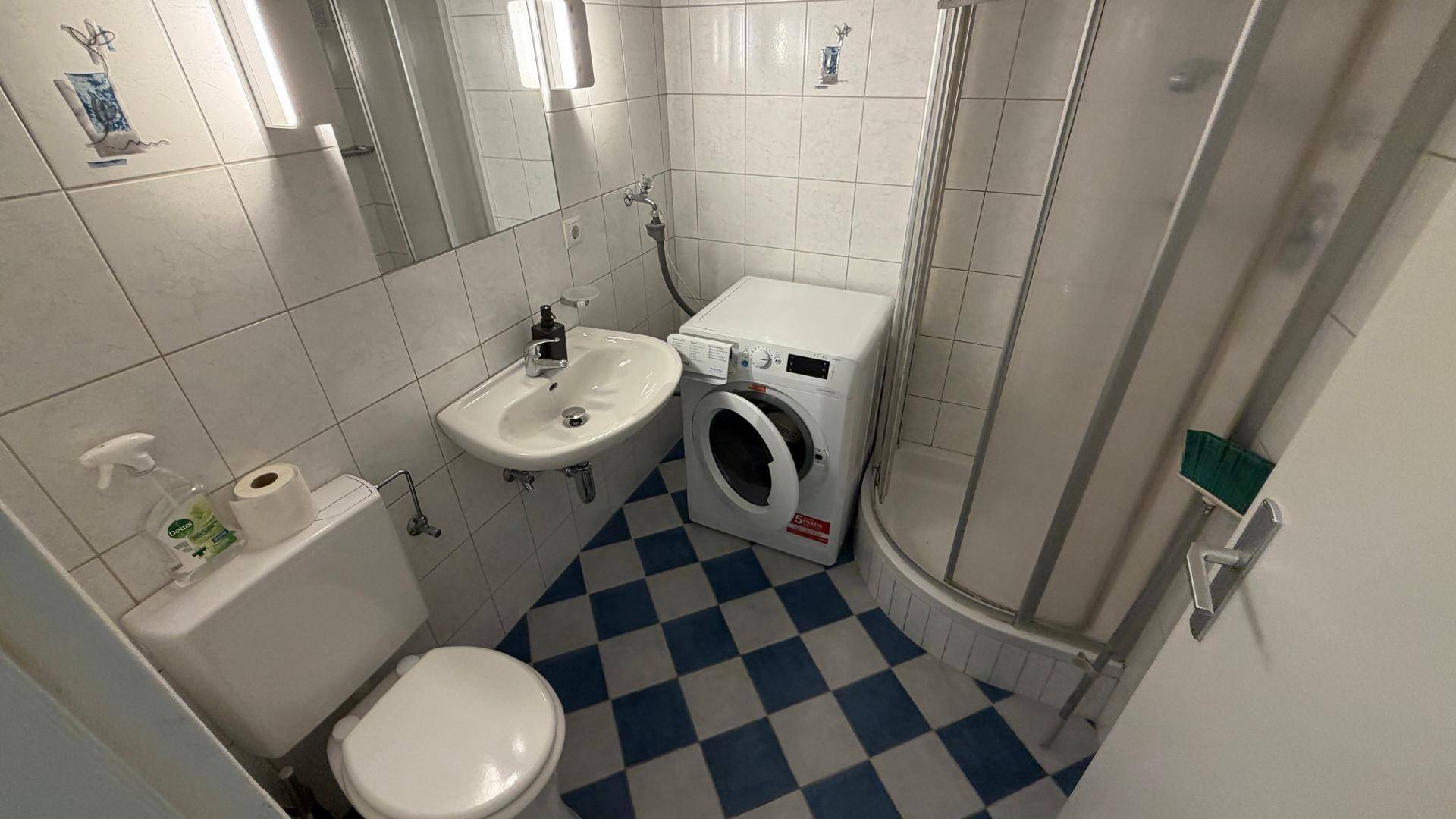 Funktionales Badezimmer mit Dusche, Waschbecken, WC und einer Waschmaschine auf kariertem Fliesenboden.