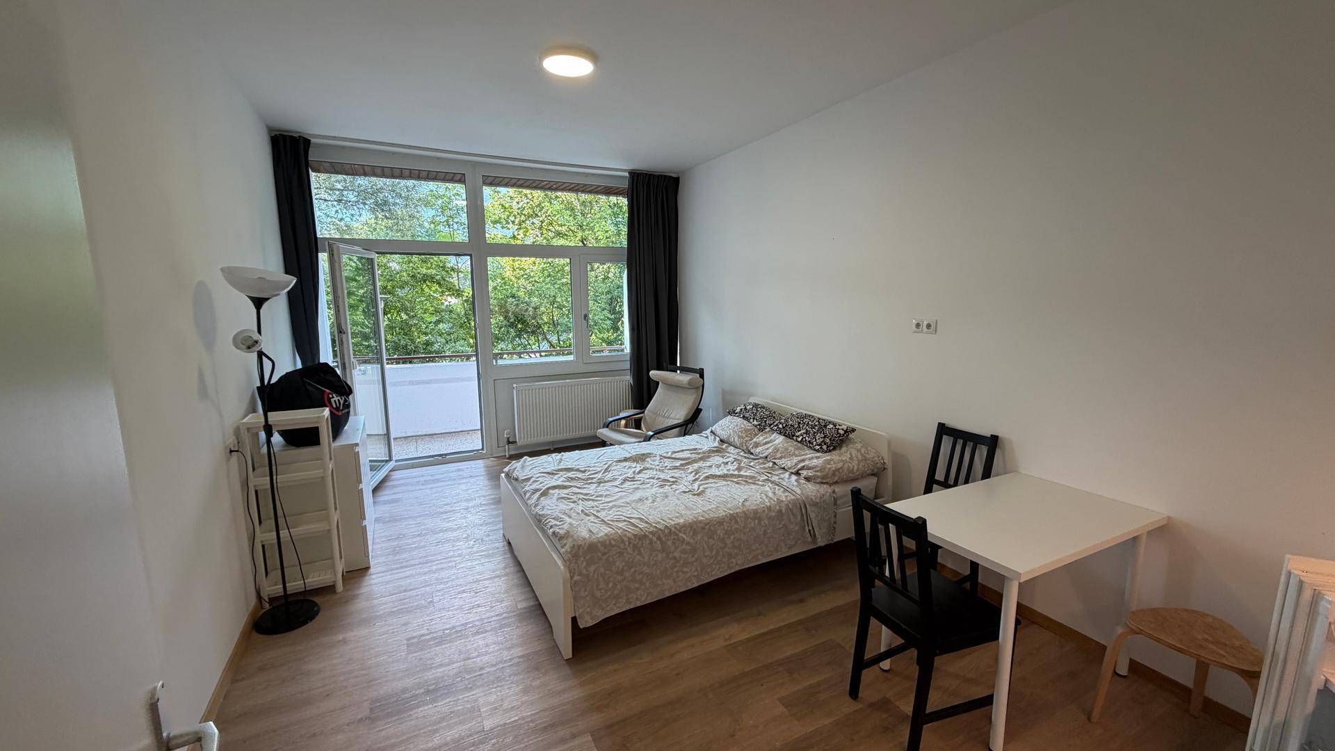 Geräumiges Studiozimmer mit Bett, Esstisch, Holzboden und Zugang zu einem sonnigen Balkon.