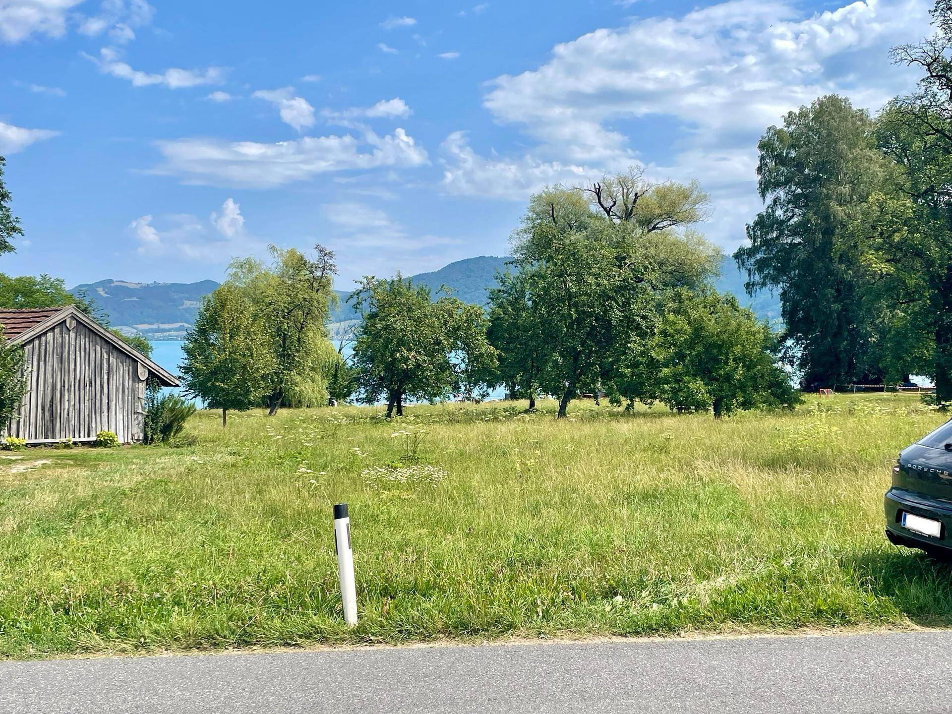 Grüne Wiese mit einem Holzschuppen und Bäumen, mit Blick auf den See und eine angrenzende Straße.