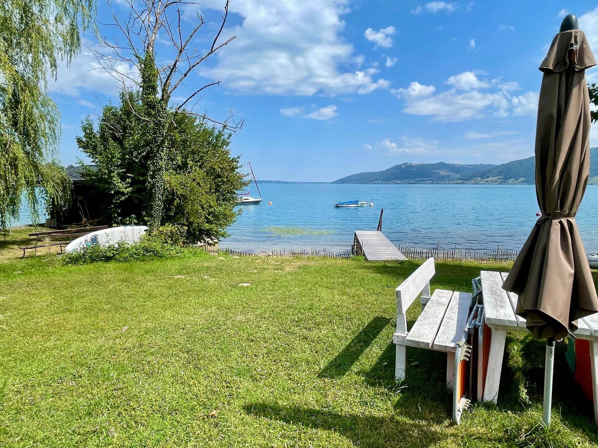 Gartenbereich mit Esstisch und Sonnenschirm, mit Blick auf den See, den Holzsteg und Boote.