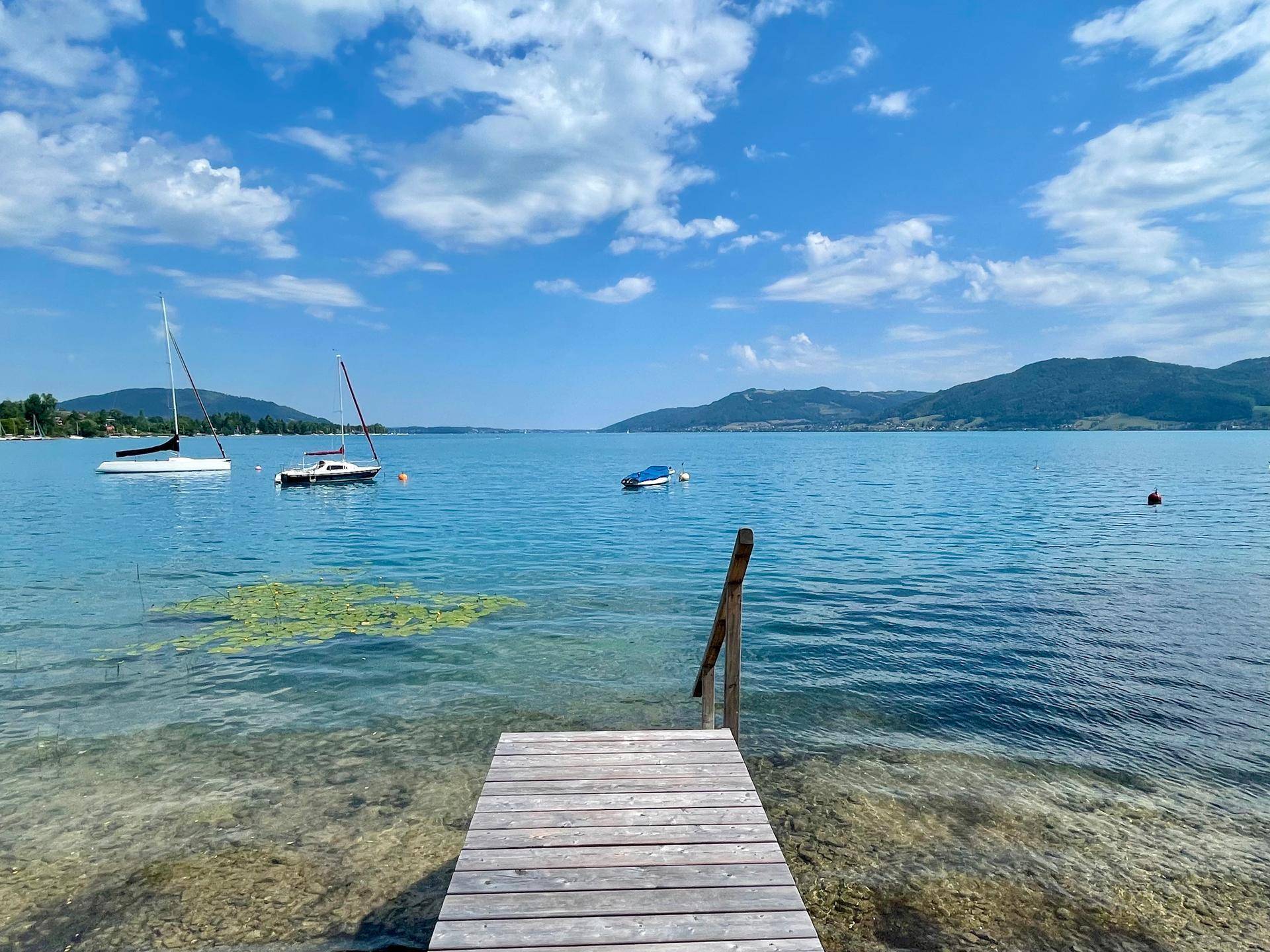 Langer Holzsteg führt ins klare Seewasser mit Segelbooten und Bergpanorama unter blauem Himmel.