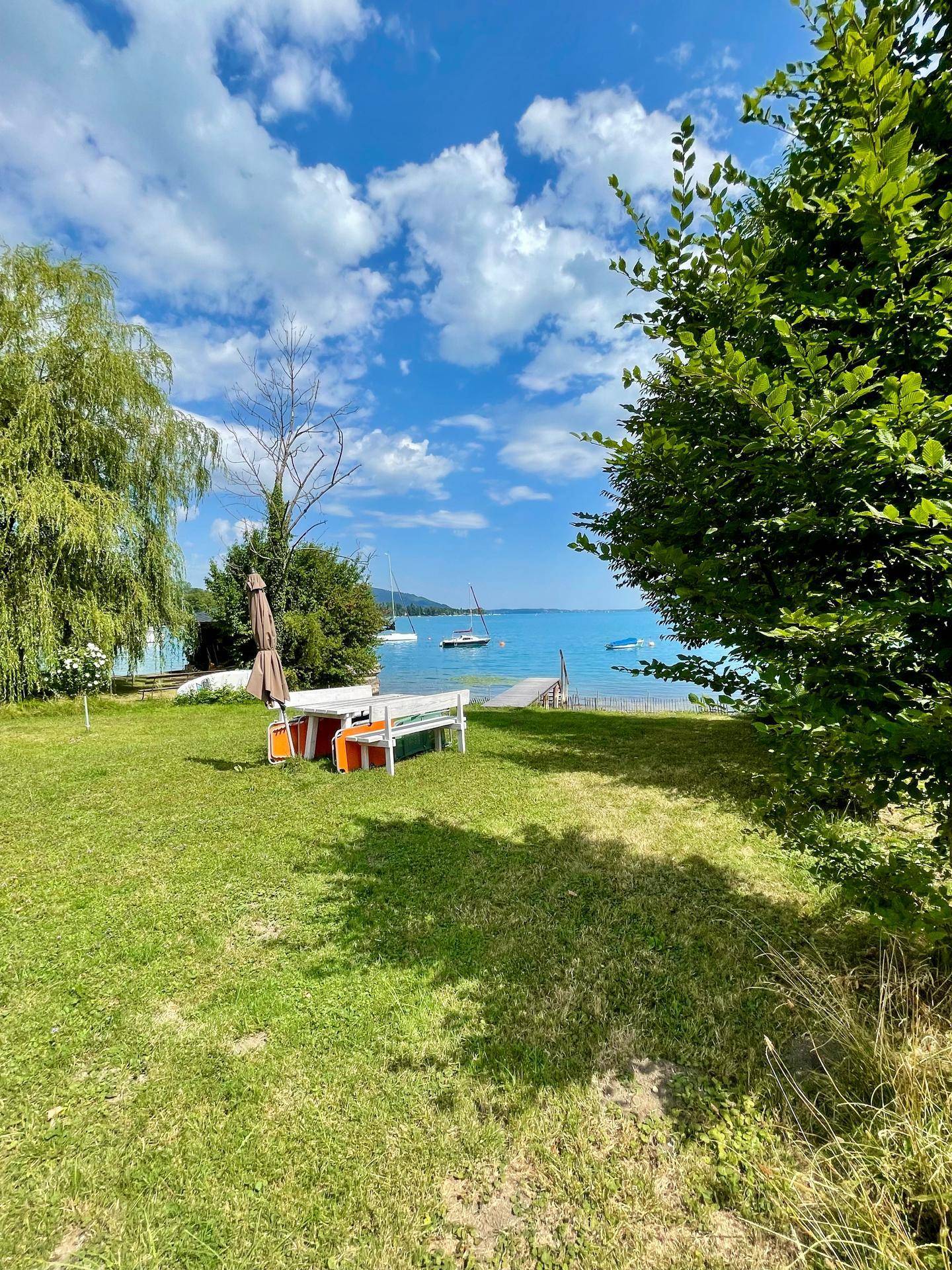 Grüne Gartenfläche mit Esstisch und Sonnenschirm, mit Blick auf den See und die Berge.