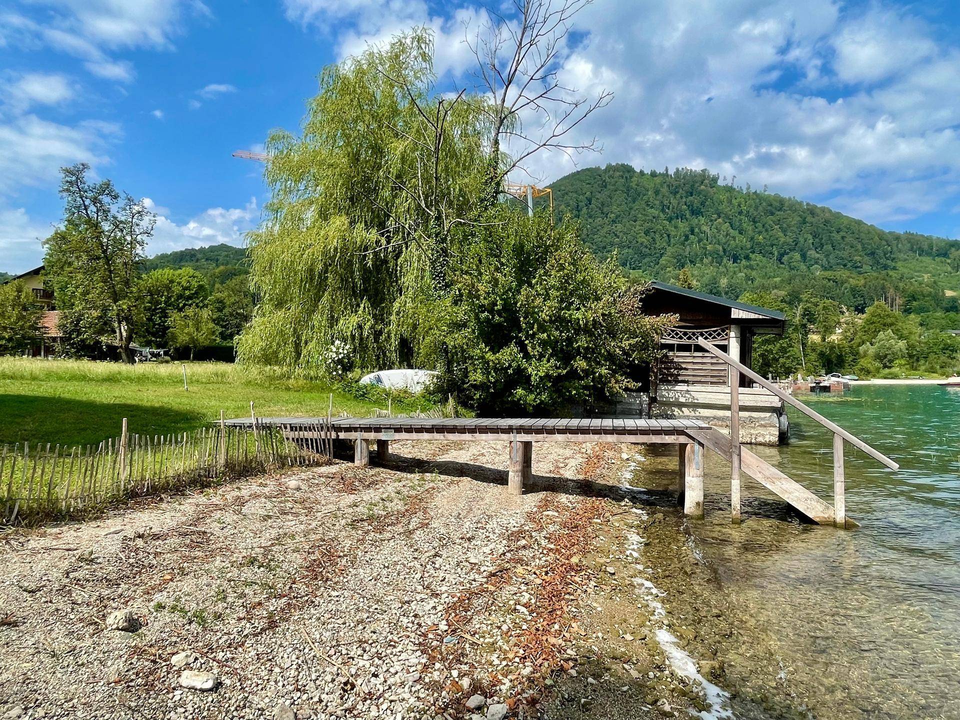 Holzsteg und Bootshaus am Seeufer, umgeben von grüner Vegetation und Bergen im Hintergrund.
