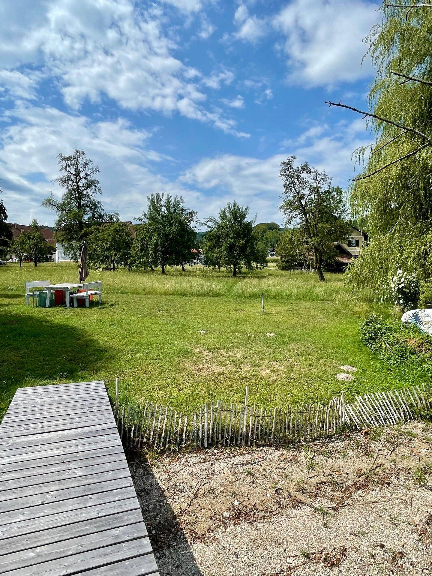 Weitläufige grüne Wiese mit Bäumen und einer weißen Sitzgruppe, vom Holzsteg aus gesehen.