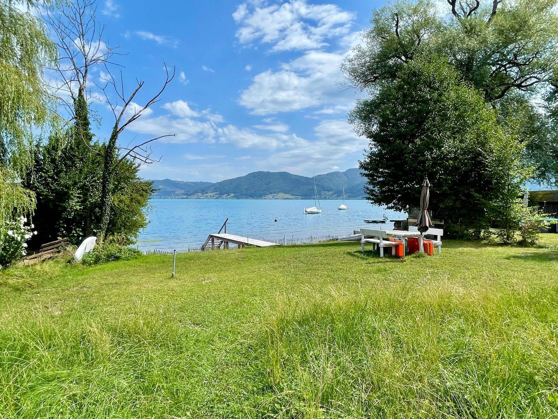 Weitläufige grüne Wiese mit Bäumen und einer Sitzgruppe, mit weitem Blick auf den See und die Berge.