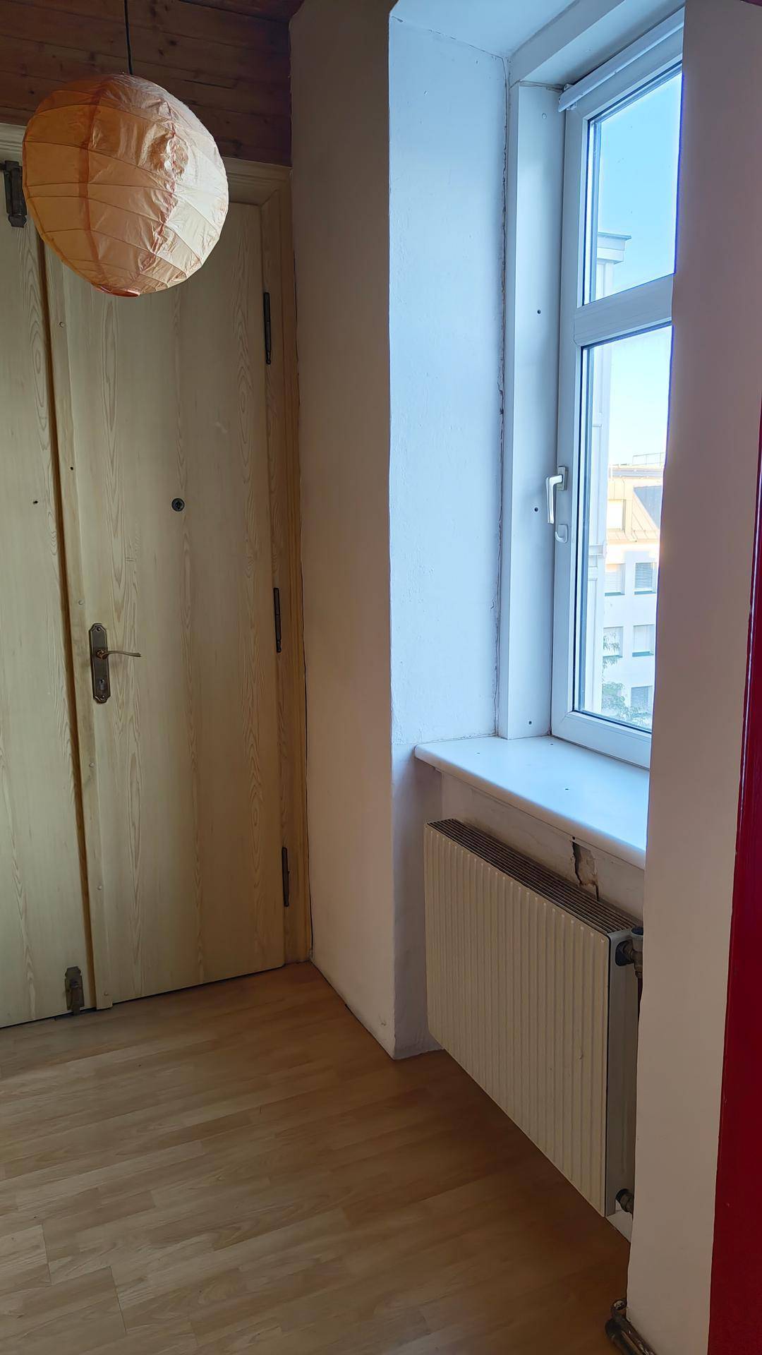 Raumecke mit hellem Holzfußboden, Fenster mit Heizkörper, Doppeltür und rotem Türrahmen.