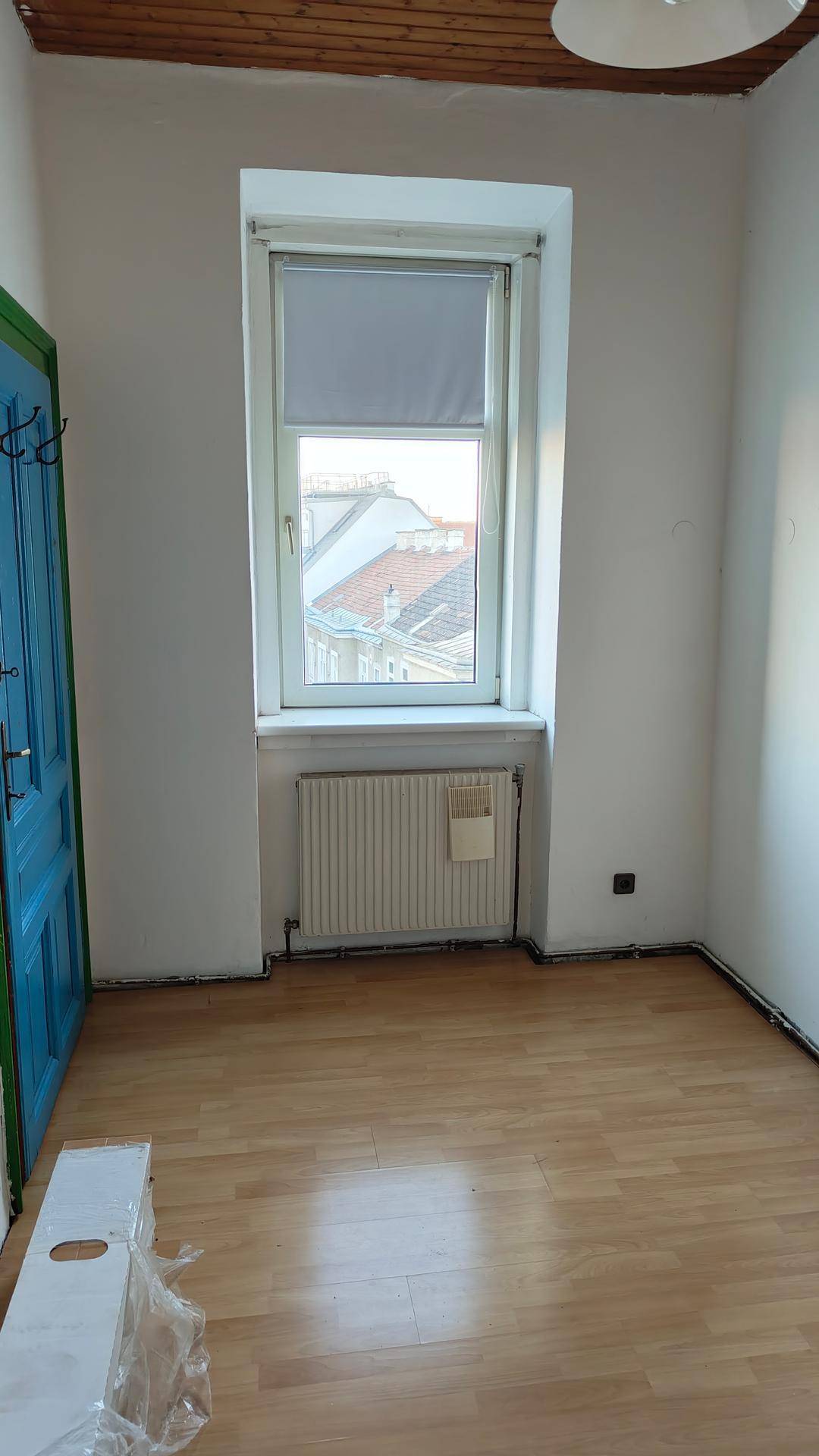 Leerer Raum mit hellem Laminatboden, weißen Wänden, Fenster mit Heizkörper und blauem Türrahmen.