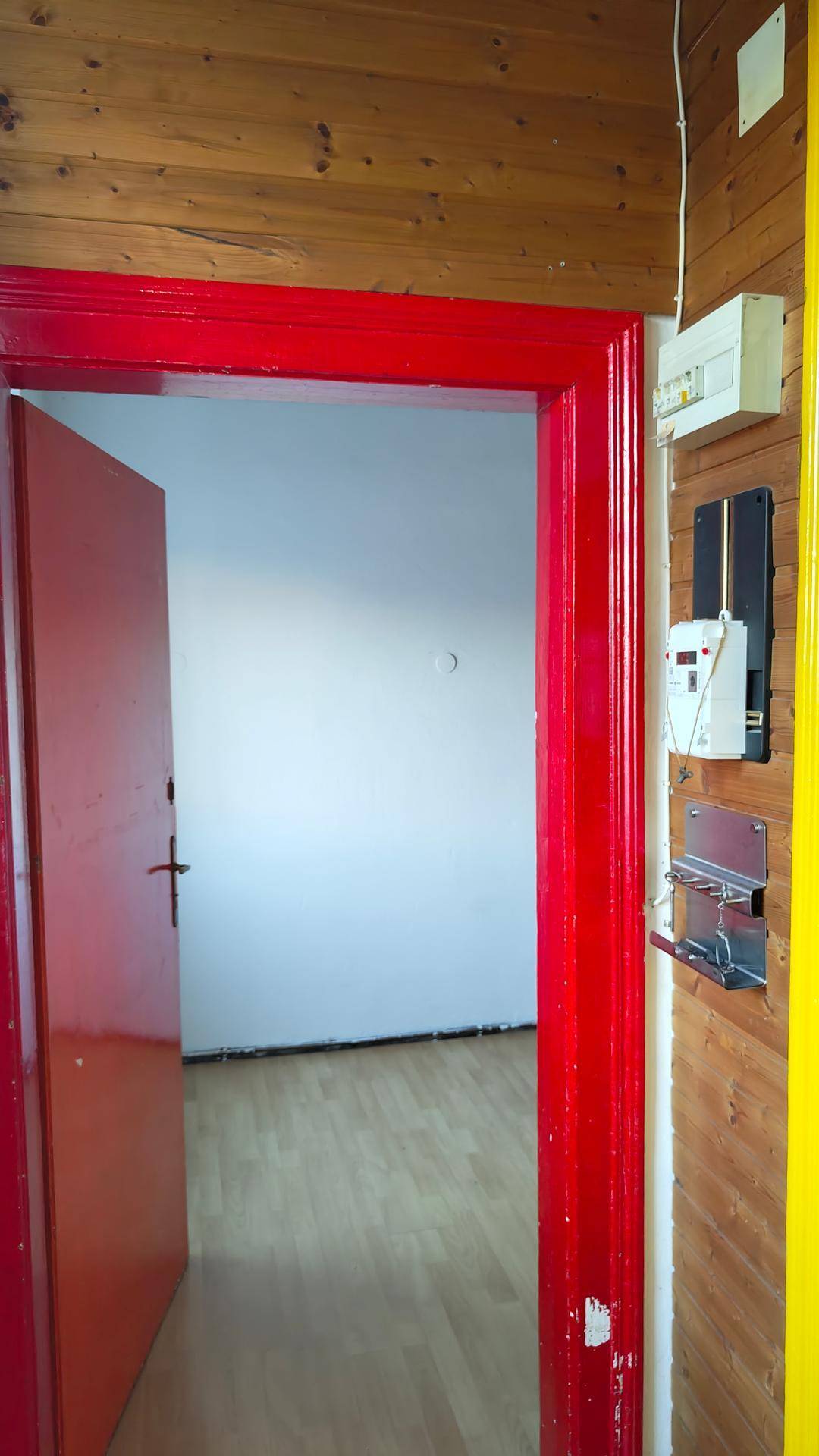 Blick durch einen roten Türrahmen in einen leeren Raum mit hellem Holzfußboden und weißen Wänden.