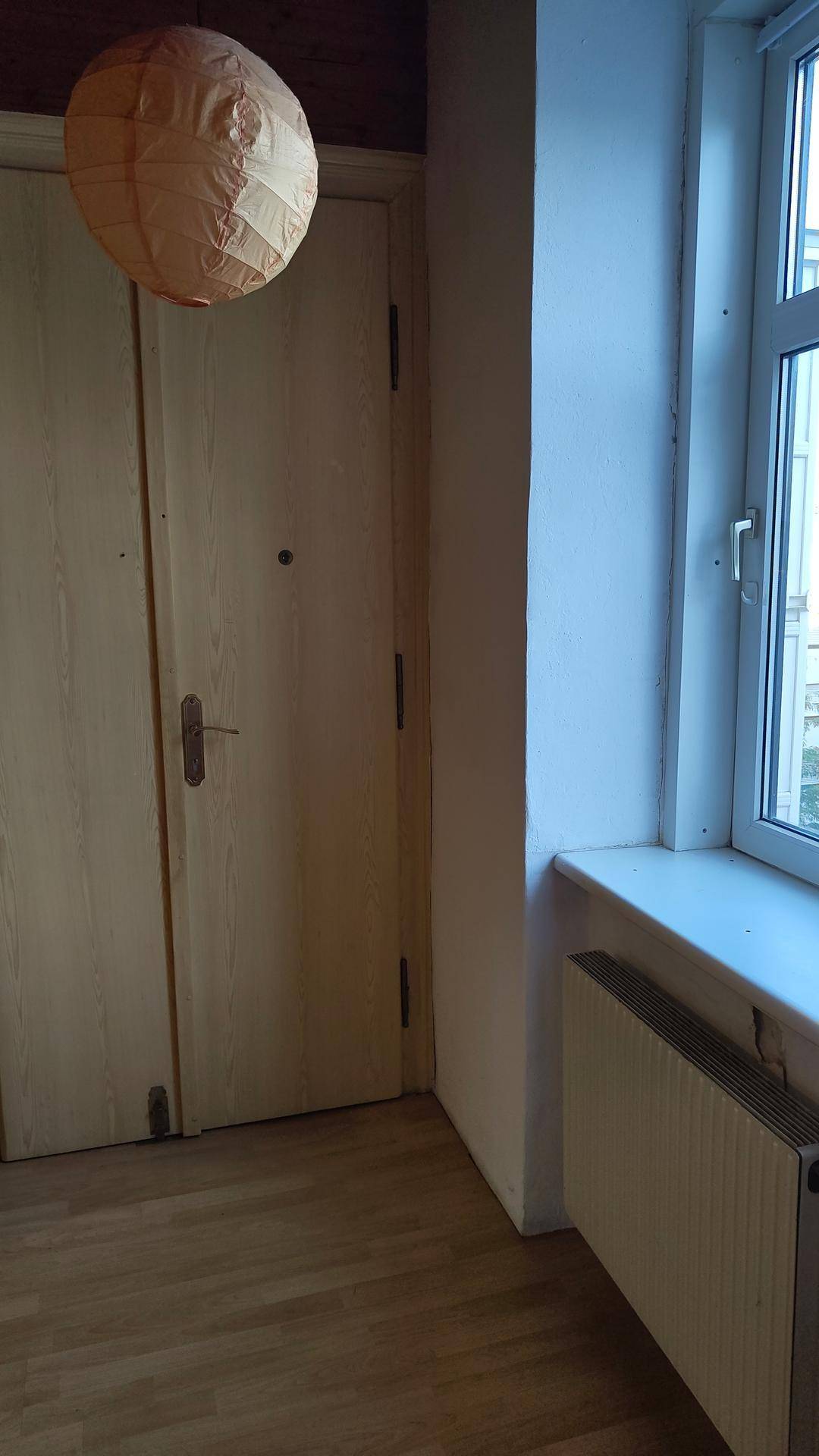 Raumecke mit hellem Holzfußboden, Fenster mit Heizkörper und einer Doppeltür, beleuchtet von einer Laterne.