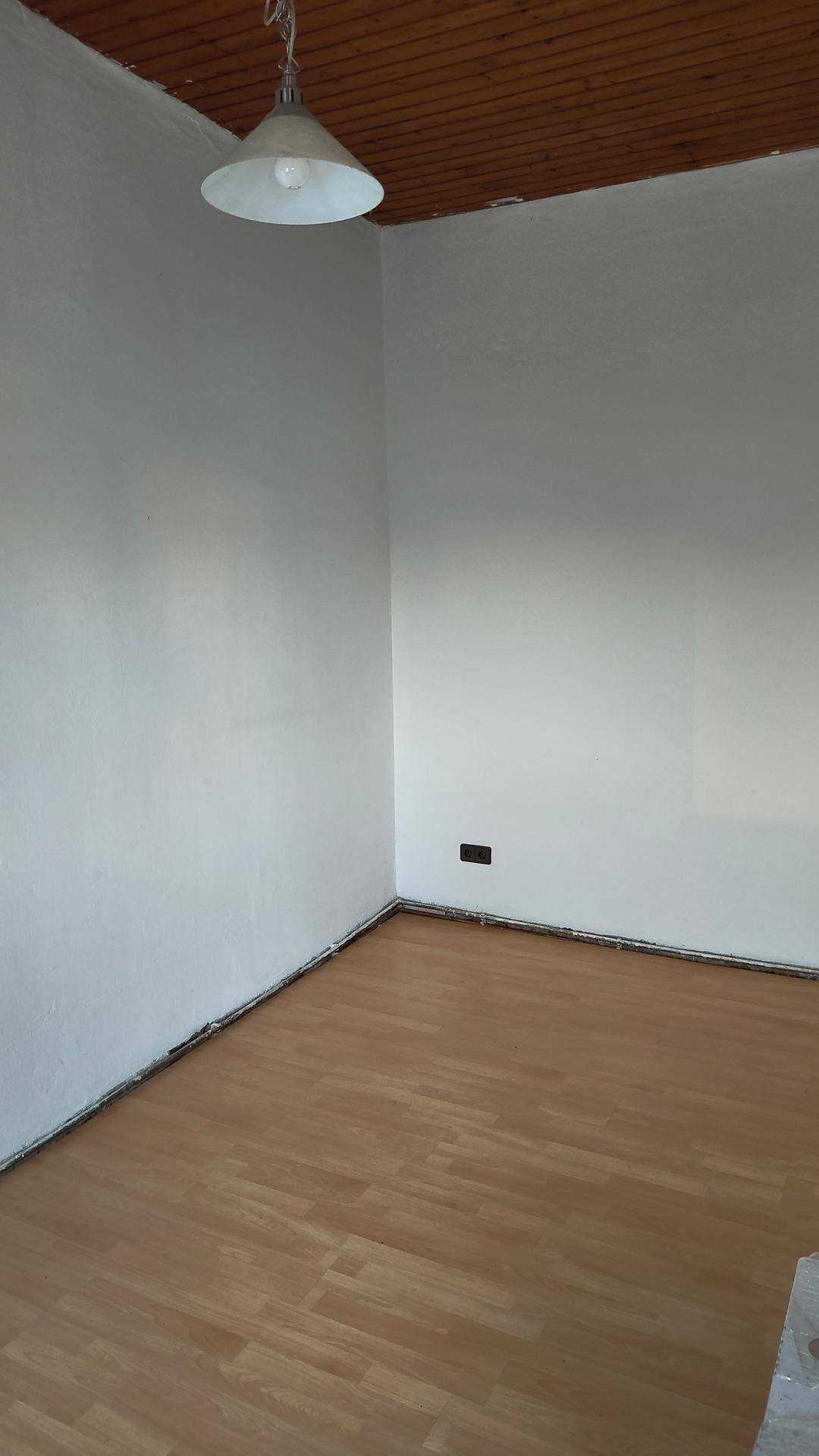 Leerer Raum mit hellem Laminatboden, weißen Wänden und einer Holzdecke mit einfacher Pendelleuchte.