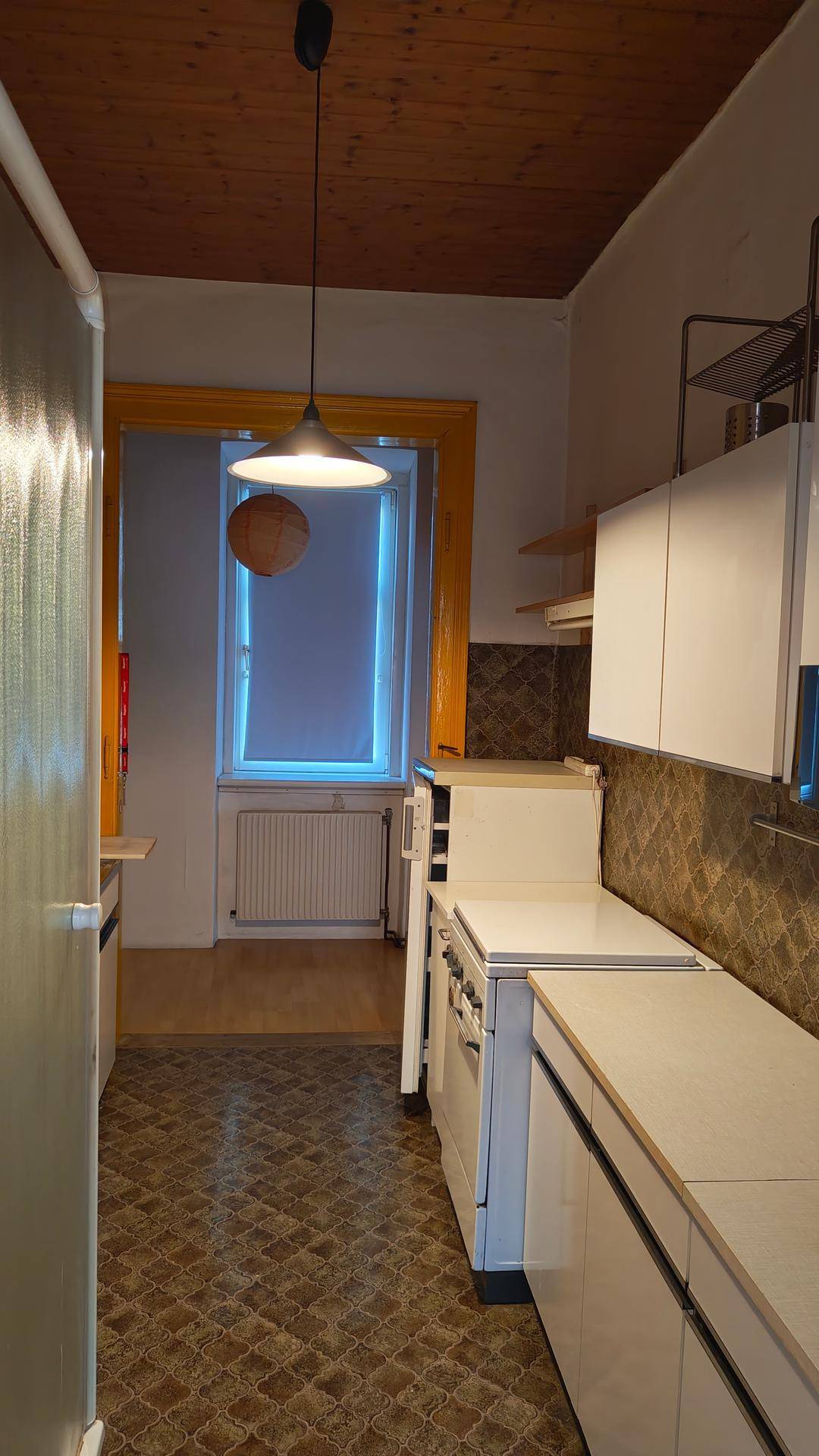 Schmale Küche mit gemusterten Fliesen, weißen Schränken, Herd und Kühlschrank, mit Holzdecke.