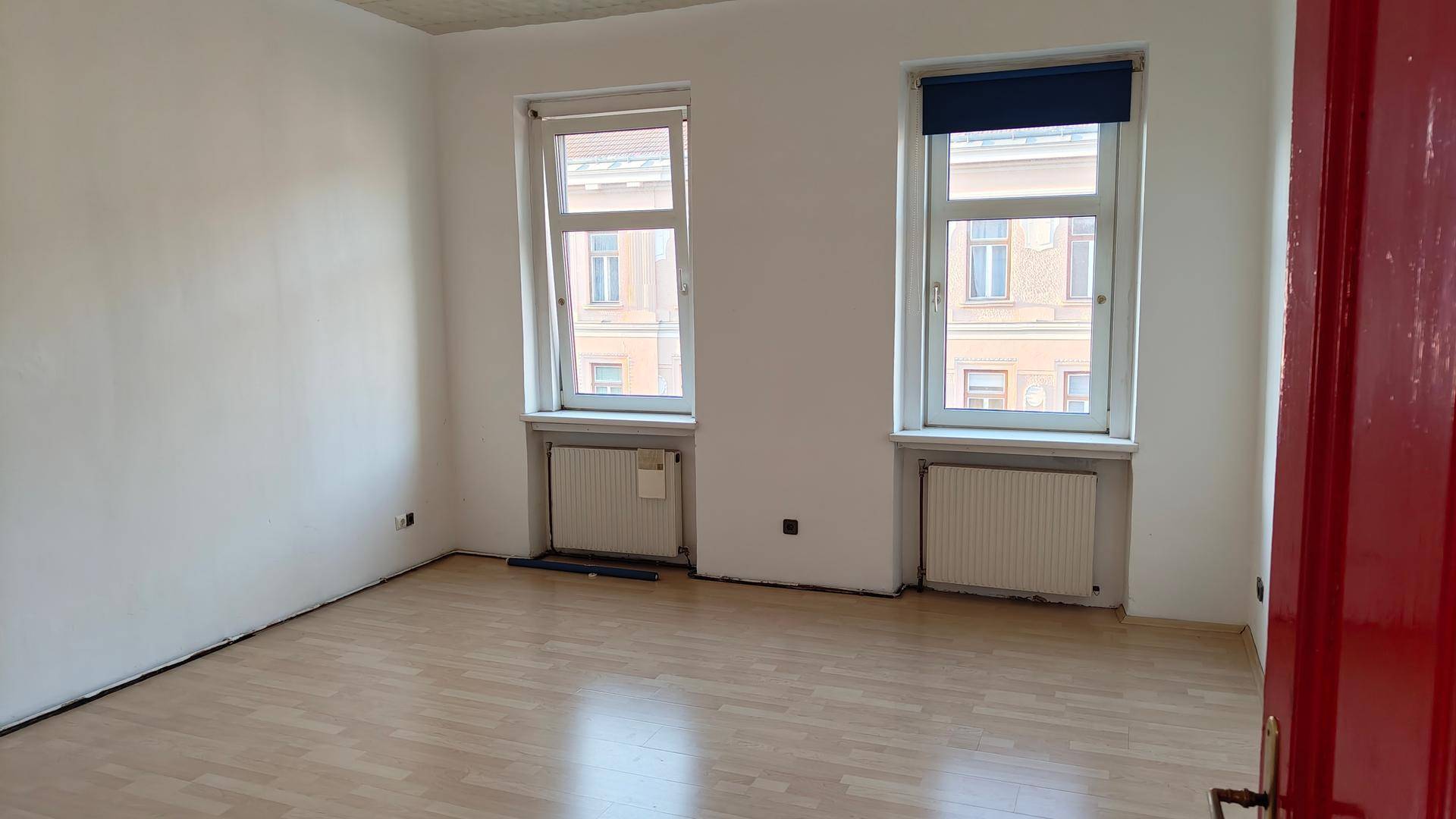 Leerer Raum mit hellem Laminatboden, weißen Wänden und zwei Fenstern mit Heizkörpern darunter.