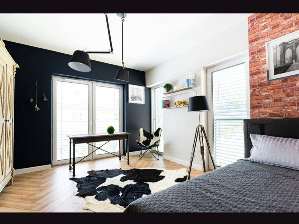Vielseitiges Zimmer mit Bett, Schreibtisch, freiliegender Ziegelwand und großen Fenstern.