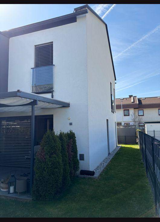 Moderne Hausfassade mit weißem Putz, kleinen Grünflächen und überdachtem Terrassenbereich.