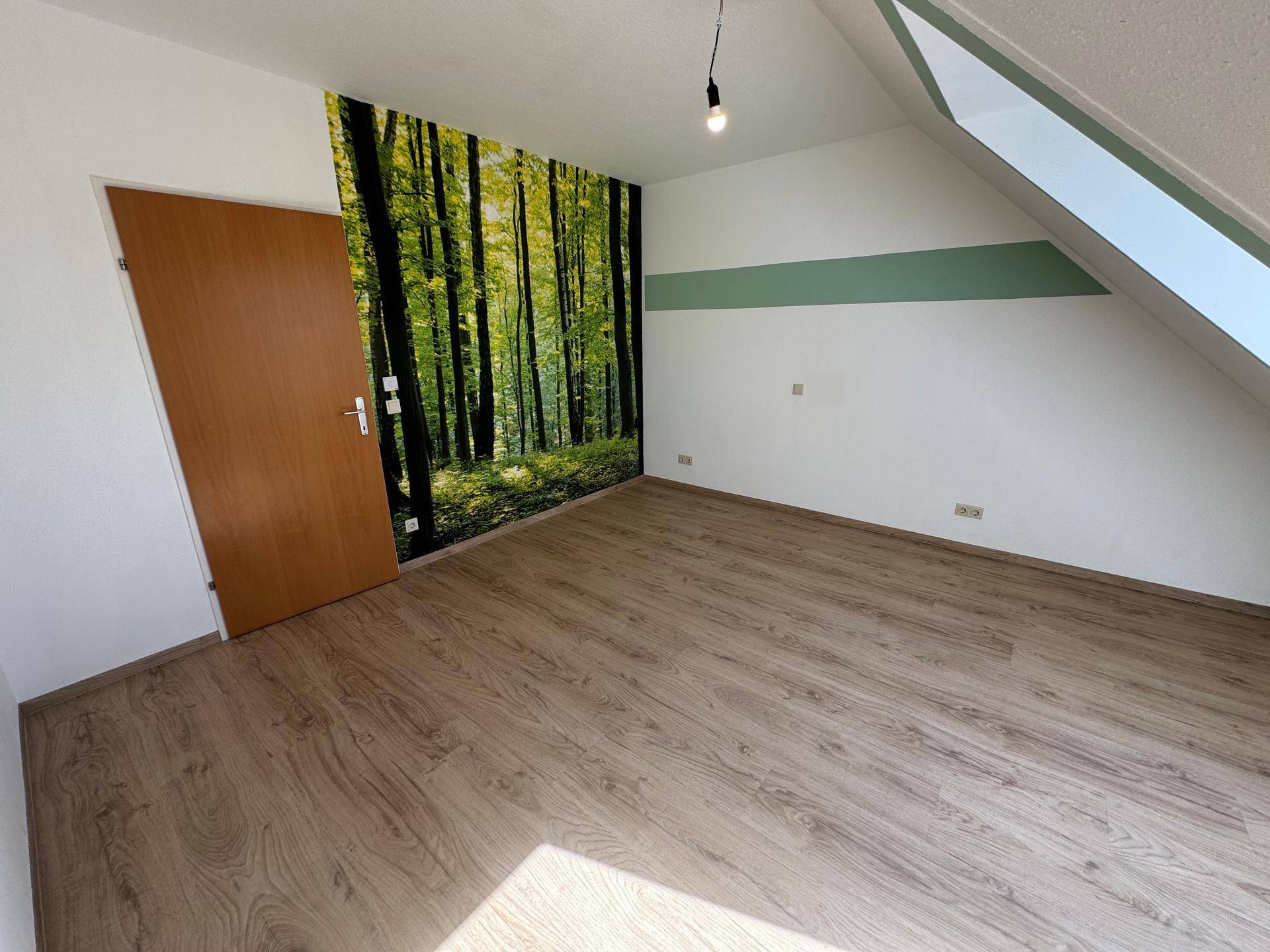 Helles Zimmer mit Laminatboden, einer Waldtapete als Akzentwand und Dachschräge.