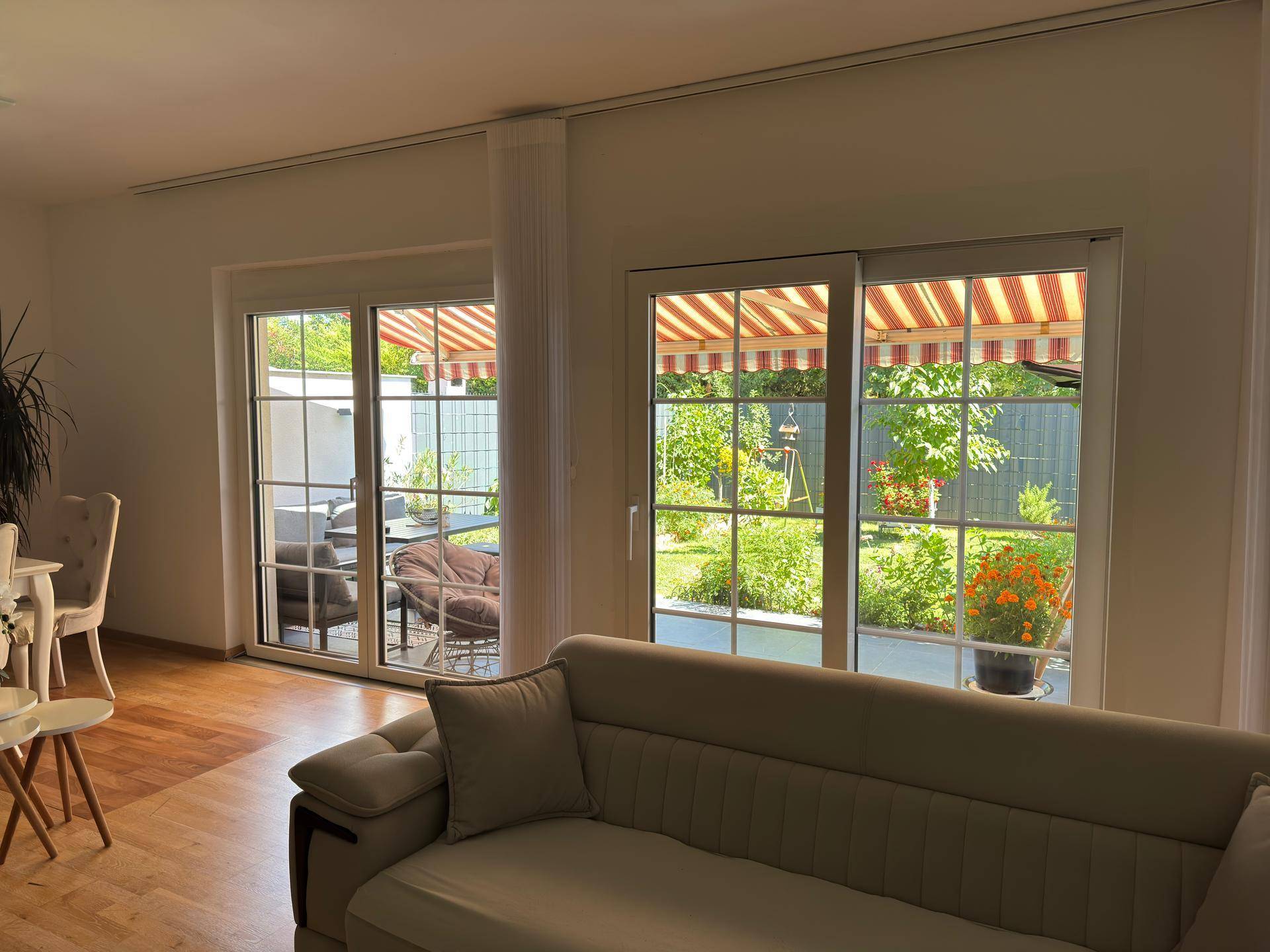 Wohnzimmer mit großen Fenstern und Blick auf die grüne Terrasse und den Garten mit Markise.