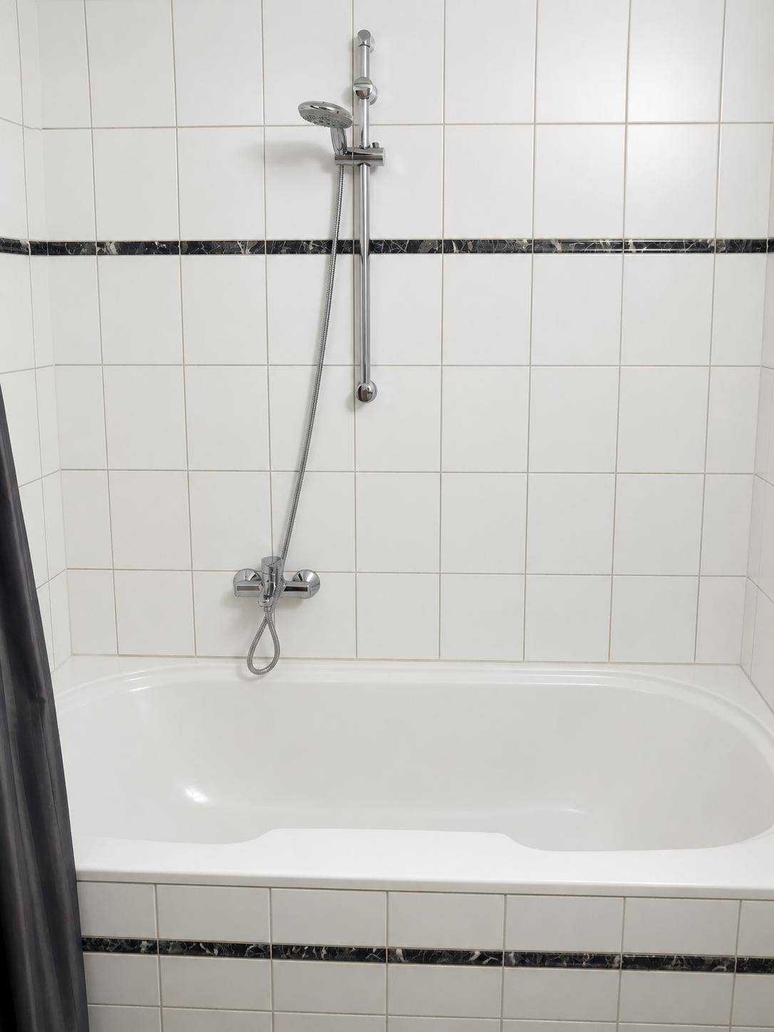 Eingebaute Badewanne mit Duscharmatur und weißen Fliesen mit schwarzem Dekorstreifen.