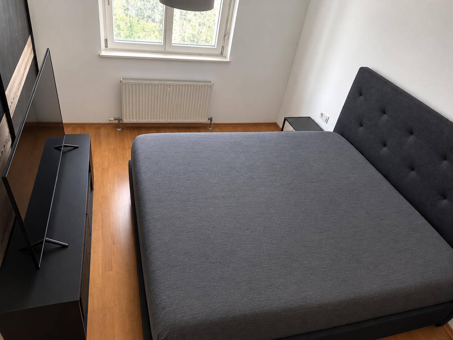 Schlafzimmer mit dunklem Doppelbett, Fernseher und hellem Laminatboden.