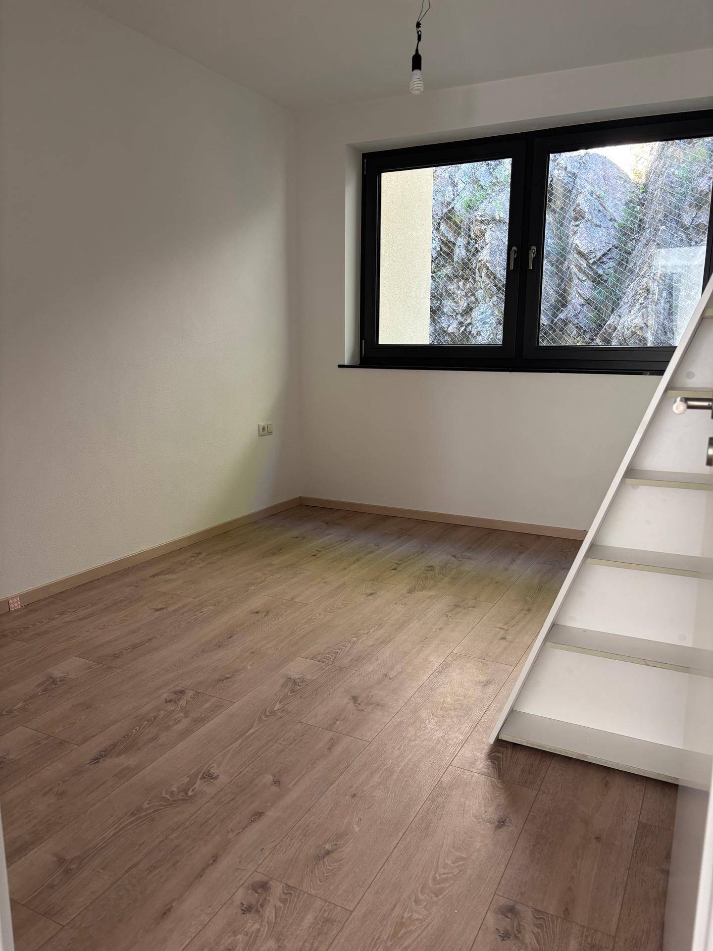 Kleines Zimmer mit Laminatboden, Fenster und einer weißen Treppe, ideal für ein Hochbett.