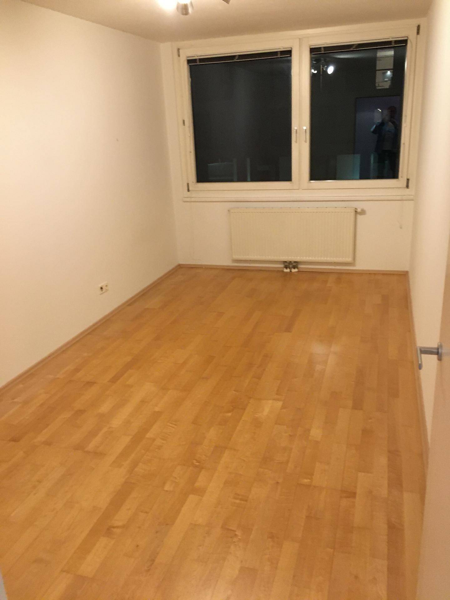 Leeres Zimmer mit hellem Holzboden, weißen Wänden und einem Fenster für natürliches Licht.