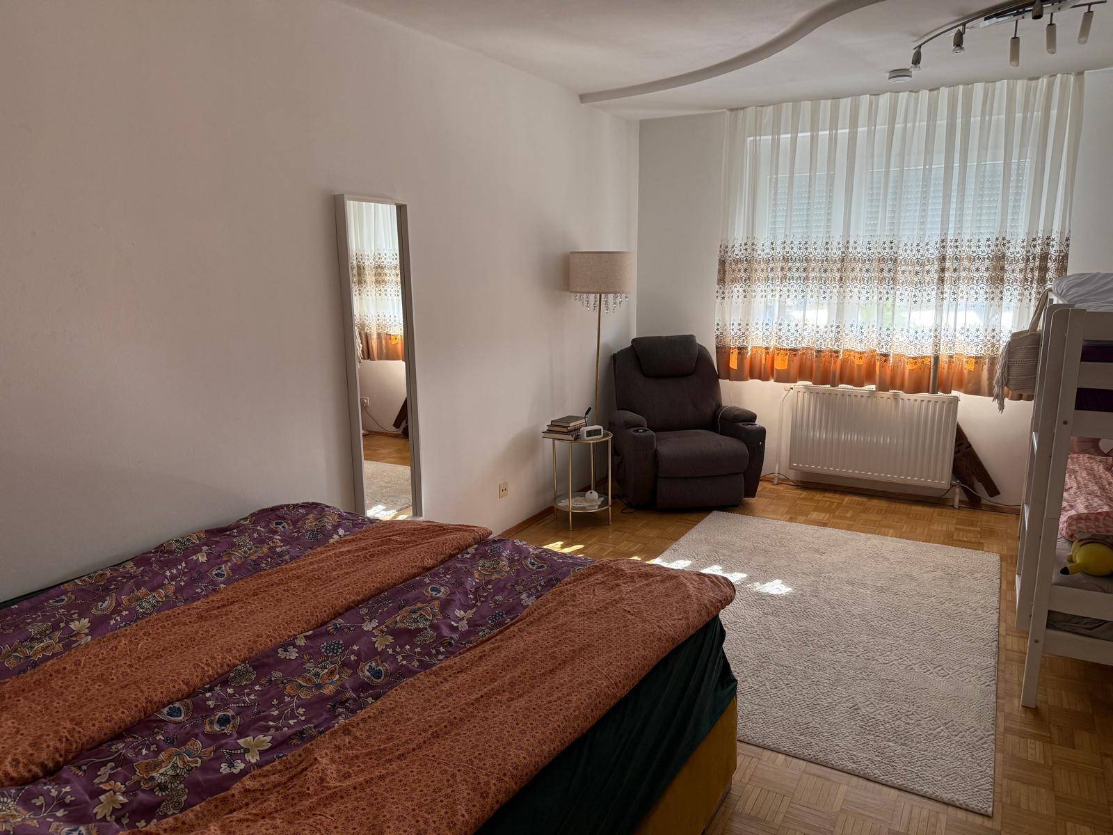 Gemütliches Schlafzimmer mit Doppelbett, Sessel und einem Etagenbett, hell durch großes Fenster.