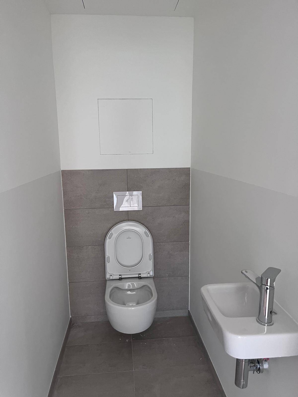 Gäste-WC mit wandhängender Toilette und kleinem Waschbecken, teilweise gefliest in Grau.