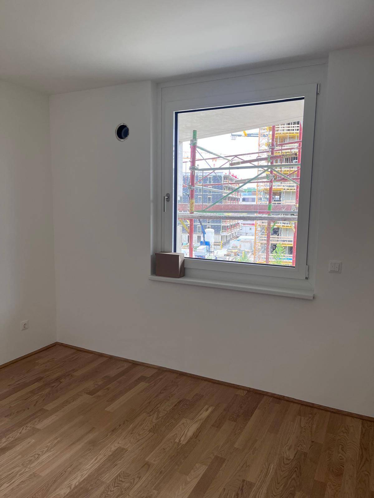 Leerer Raum mit Holzboden und einem Fenster, das einen Blick auf eine Baustelle bietet.