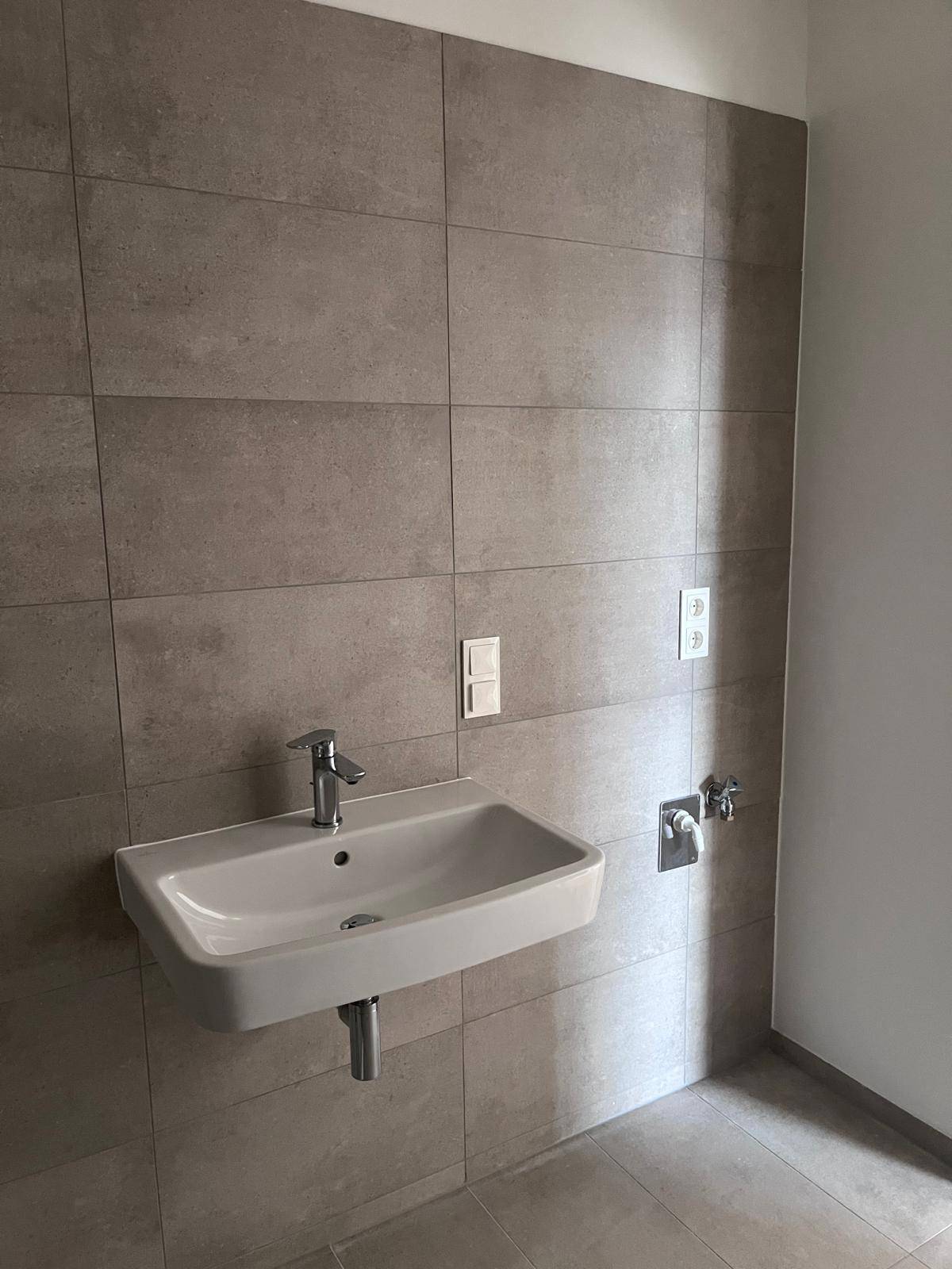 Modernes Badezimmer mit wandmontiertem Waschbecken und großformatigen grauen Fliesen an Wänden und Boden.