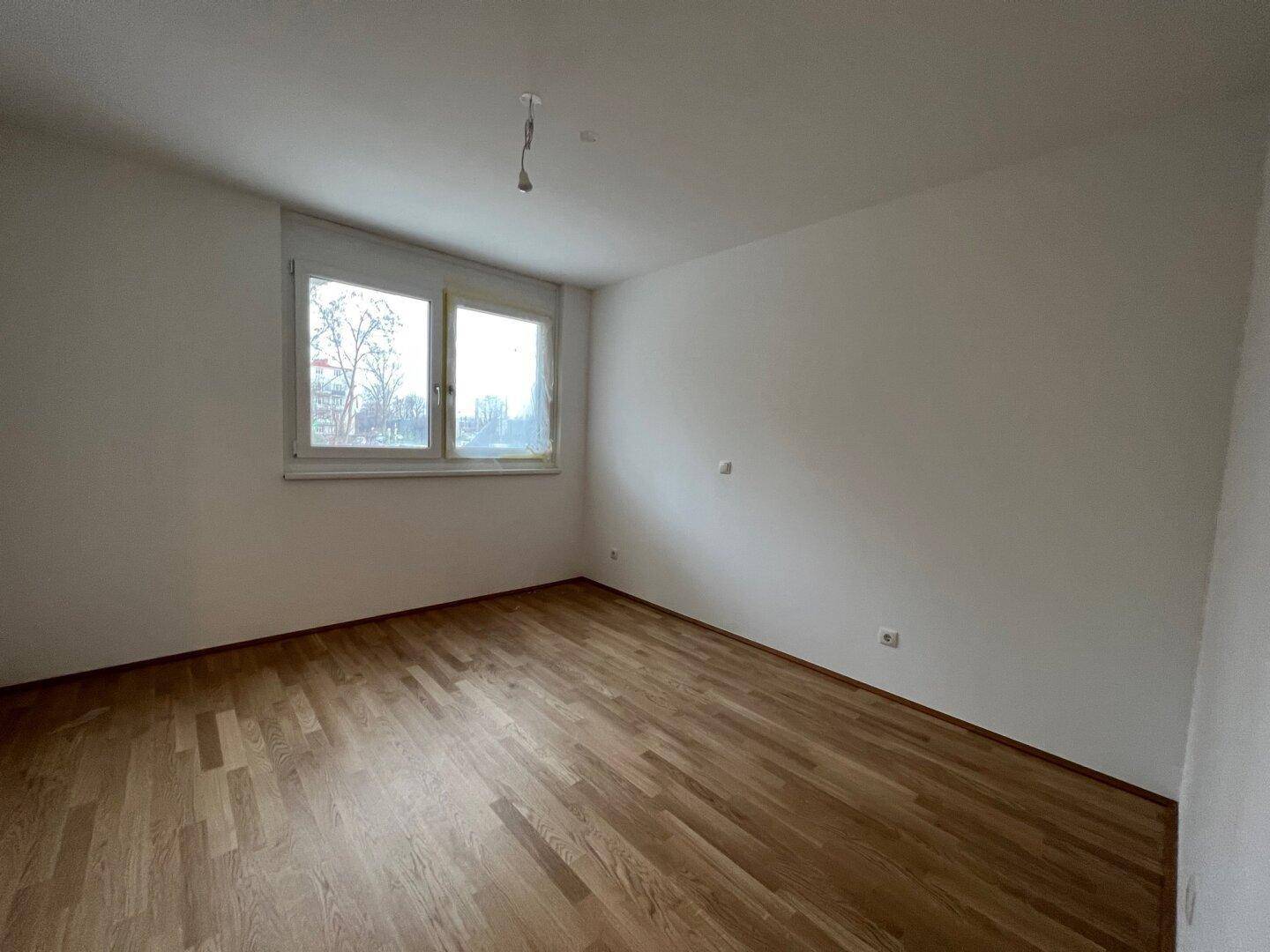 Leeres Schlafzimmer mit Parkettboden, weißen Wänden und einem großen Fenster.